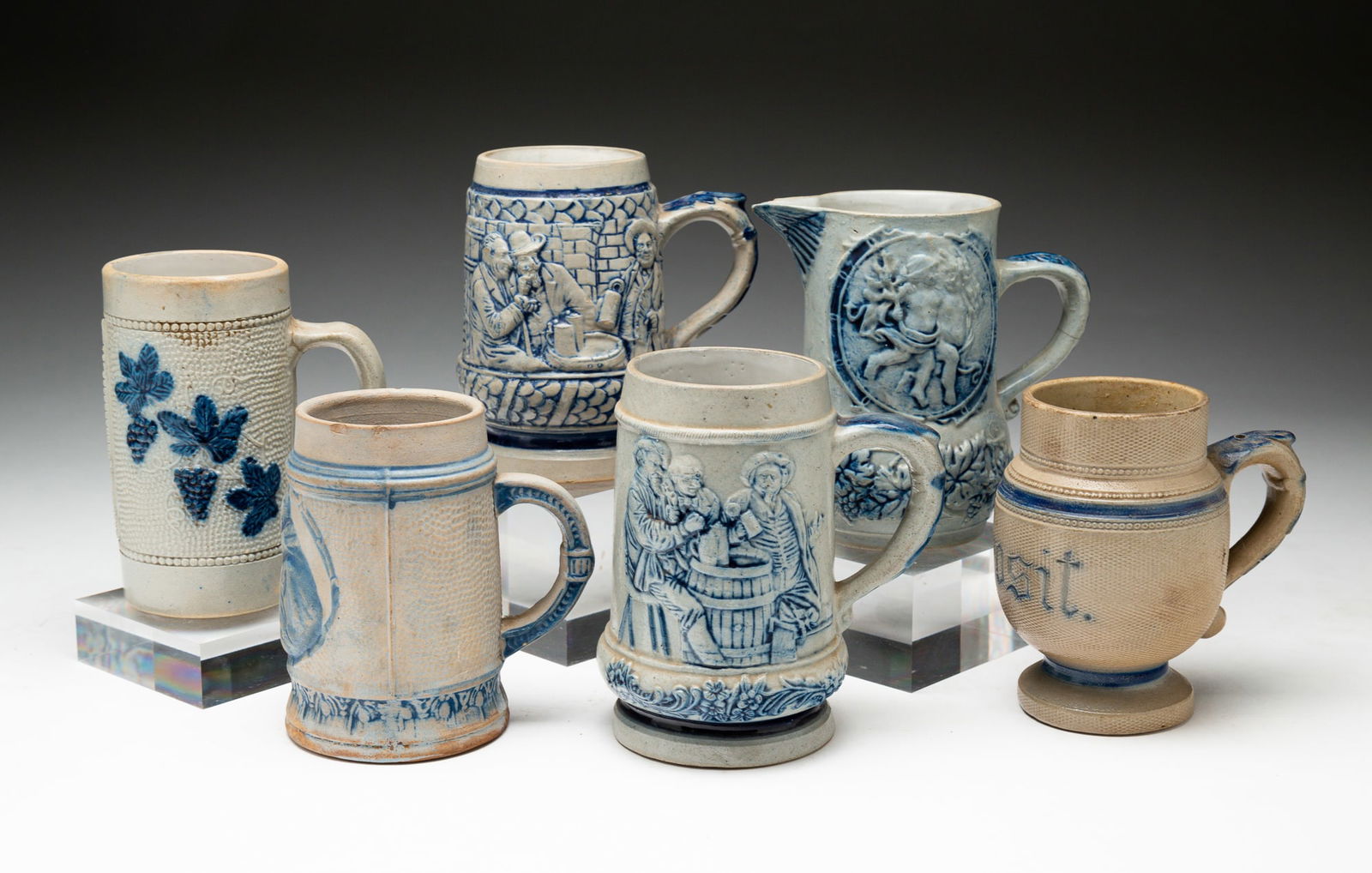 FIVE BLUE AND GRAY SALT-GLAZED STONEWARE STEINS AND A PITCHER.: American, late-19th to early-20th Century. Attributed to Whites Pottery, Utica, NY. Six pieces total. The first stein embossed "Prosit." Repaired crack to handle. 5"h. The second embossed "Prosit" wit