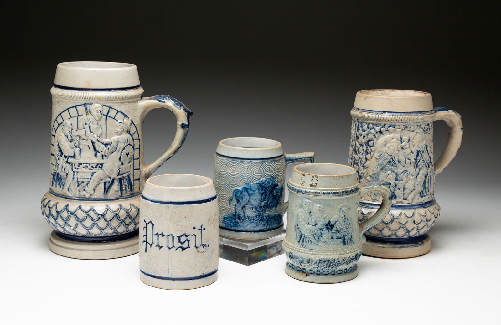 FIVE BLUE AND GRAY SALT-GLAZED STONEWARE STEINS.: American, late-19th to early-20th Century. Attributed to Whites Pottery, Utica, NY. The first molded with a tavern scene and embossed German saying on reverse. With impressed "39" on underside. Crack