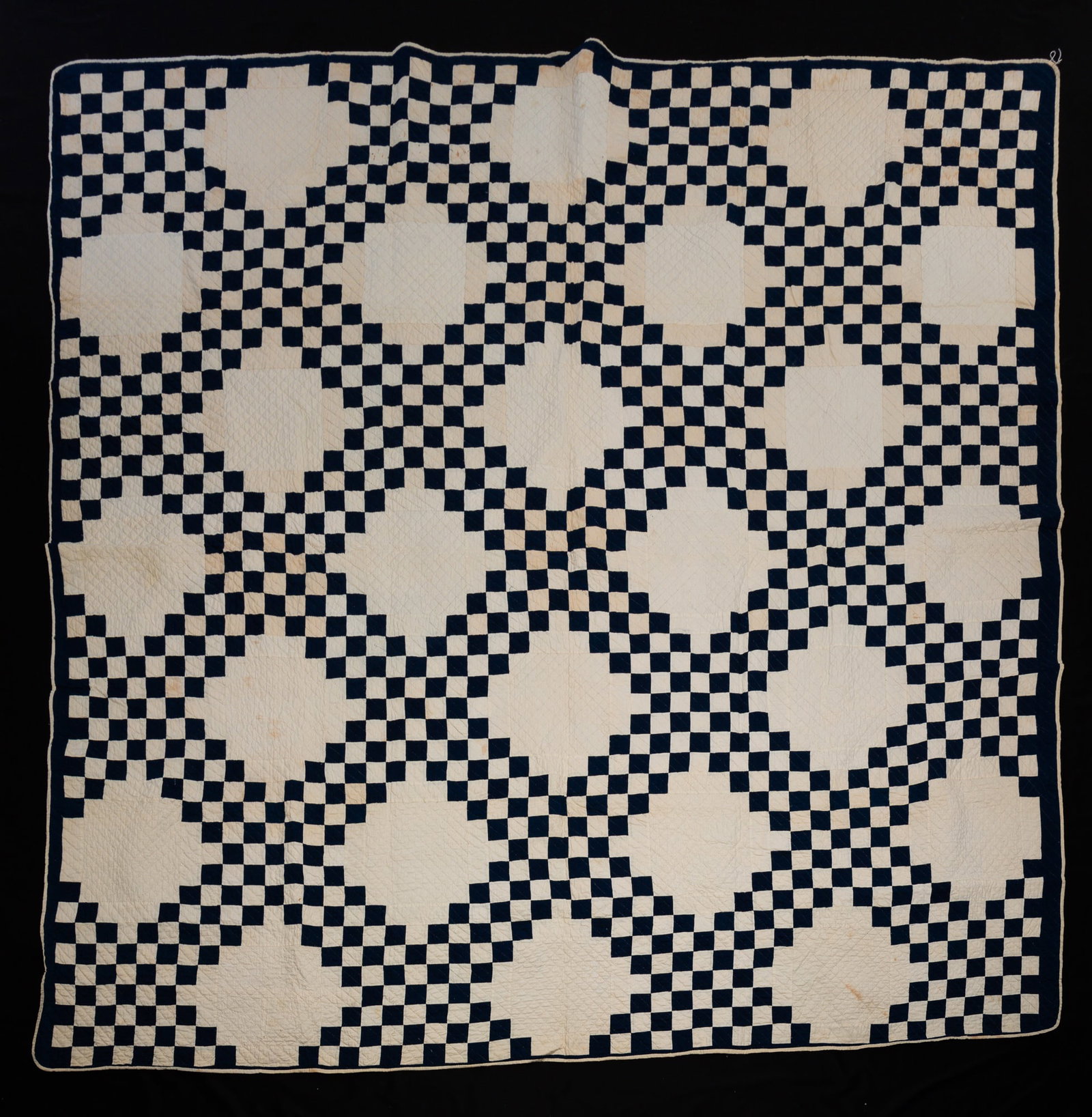 PIECEWORK QUILT IN DOUBLE IRISH CHAIN PATTERN.: American, 20th Century. Navy blue squares on an ivory ground. Staining. 84"w. 84"l.