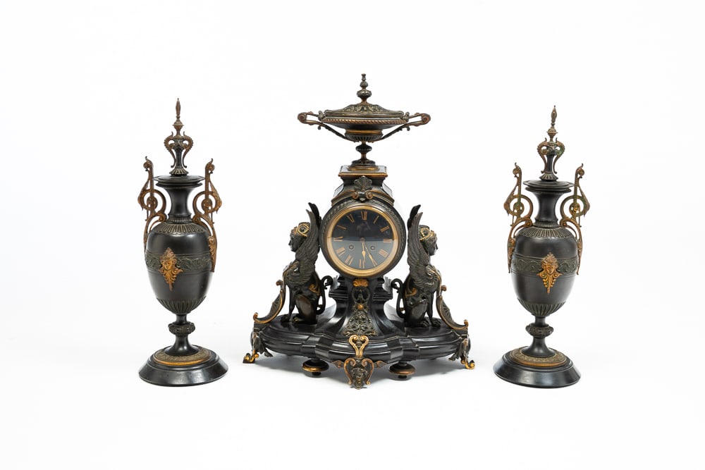 AESTHETIC PERIOD MANTLE CLOCK AND GARNITURE.: French, fourth quarter 19th Century. Slate base and central clock pier surmounted with a bronze lidded urn, flanked by bronze Greek Sphinx. Elaborate bronze ormolu, black face and Japy Frere's brass m