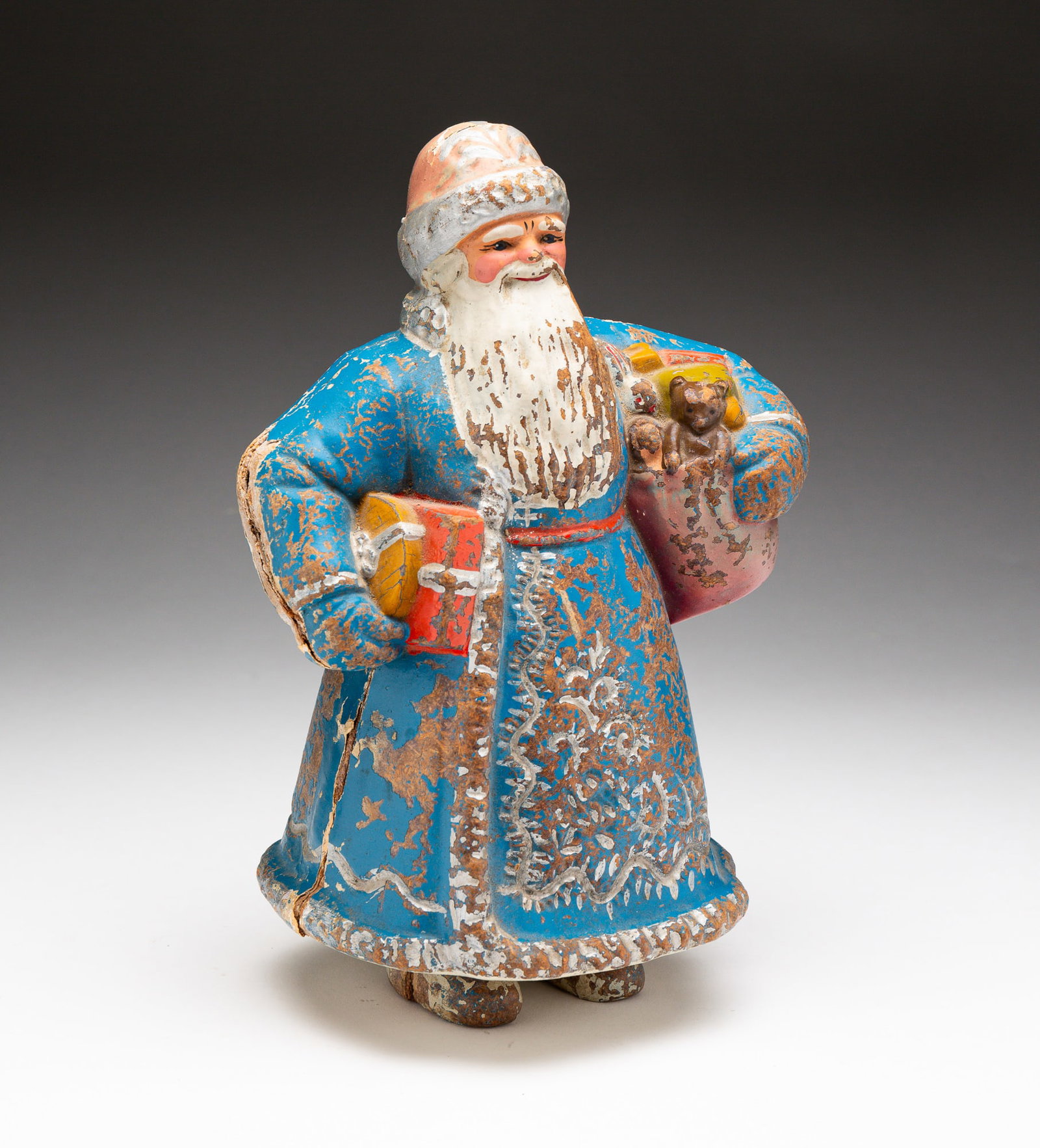 COMPOSITION FATHER CHRISTMAS IN BLUE ROBE.: Continental, late-19th or early-20th Century. Santa figure holding a sack of toys in one arm and presents in the other. Paint loss throughout. Open seam. 10"h.