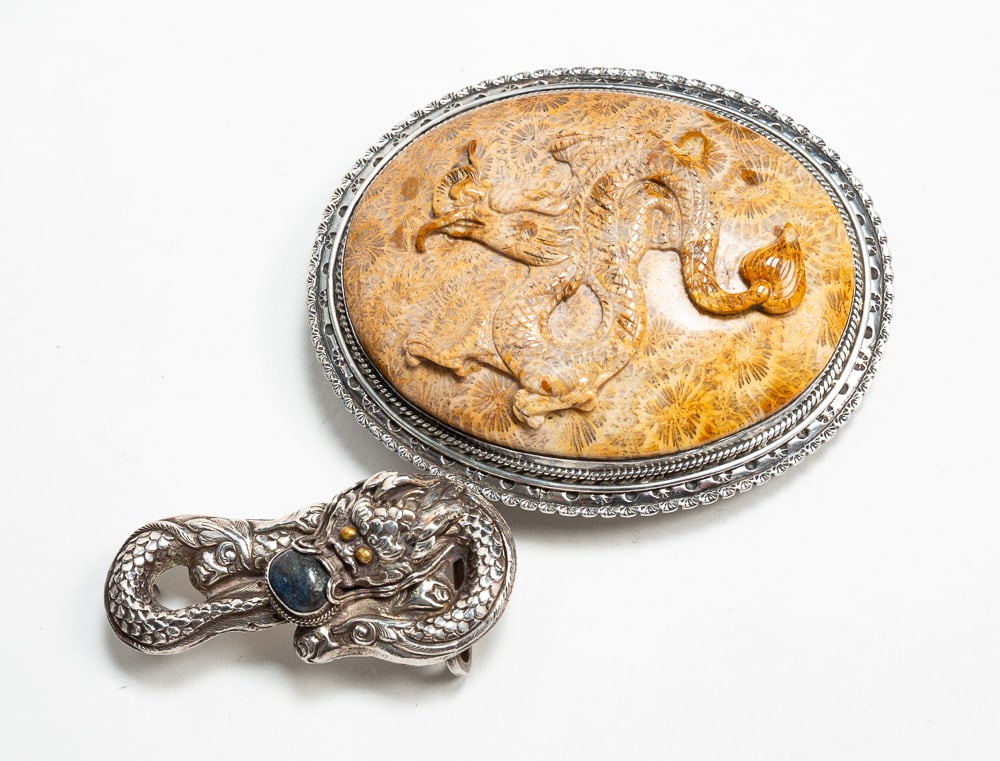 TWO DRAGON MOTIF STERLING SILVER BELT BUCKLES.: Mid- to late-20th Century. Swirling dragon with golden eyes and a bezel-set lapis lazuli stone in its mouth. Stamped "925" on back. 2.75"w. Oval shape with large bezel-set fossilized coral carved as a