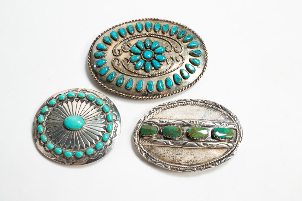 THREE SILVER BELT BUCKLES.: Mid- to late-20th Century. Oval shape with four sawtooth bezel-set turquoise stones. Marked "Sterling" with a bent arrow symbol on the back. 3.75"w. Circular shape with a central sawtooth bezel-set tu