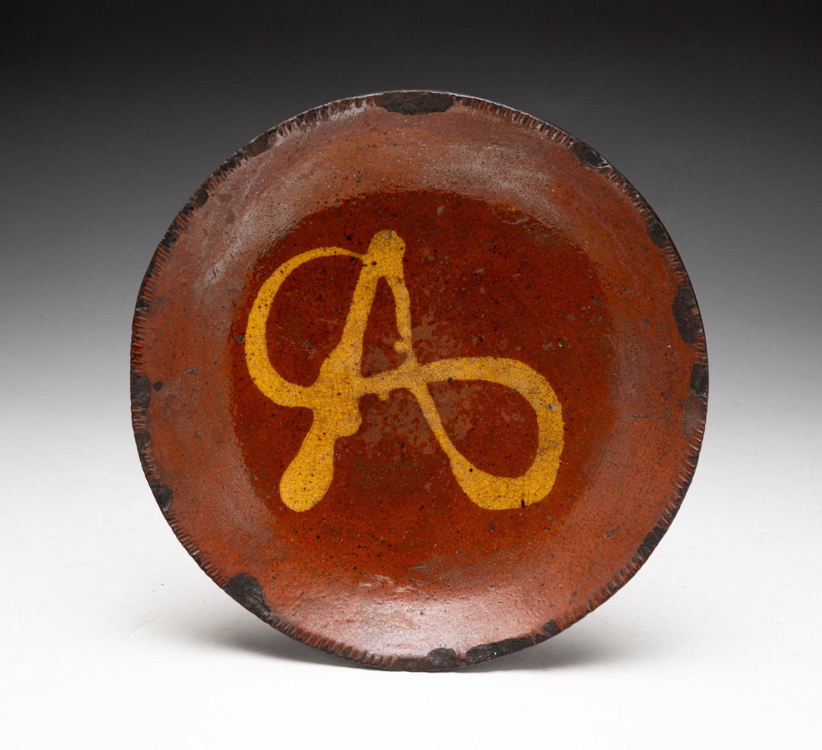 SLIP DECORATED REDWARE PLATE.: SLIP DECORATED REDWARE PLATE. American (Pennsylvania), 19th Century. Orange and black mottled glaze decorated in yellow, possibly the letter "A," with coggled rim. 8"d. From the collection of Carol Sc