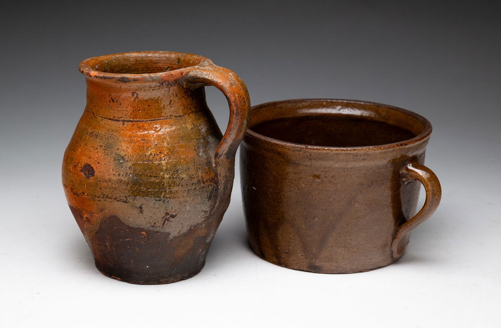TWO HANDLED REDWARE VESSELS.: TWO HANDLED REDWARE VESSELS. American, 19th Century. A squat jar with brownish-green glaze. 4"h. And an ovoid jar with orange and mottled green glaze. 6"h. From the collection of Carol Schulman (Chest