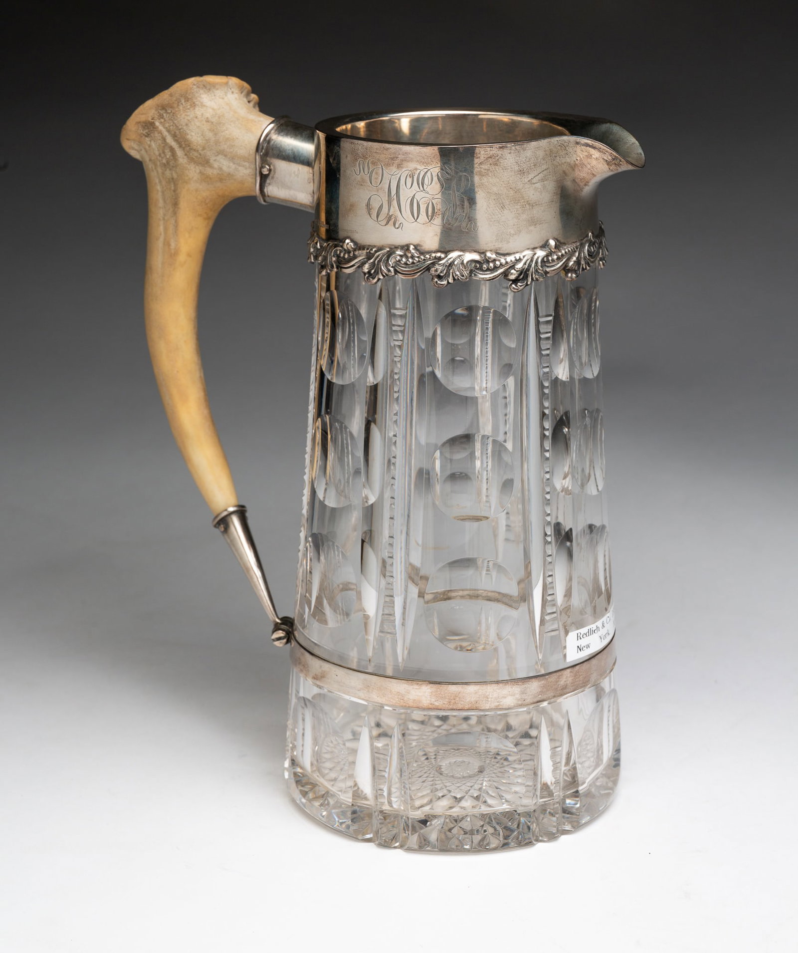 REDLICH AND COMPANY CUT GLASS PITCHER WITH STERLING MOUNTS.: American, 20th Century. Pitcher with a paneled thumbprint pattern, sterling banding and antler handle. Loring Andrews retail mark. 9.25"h.