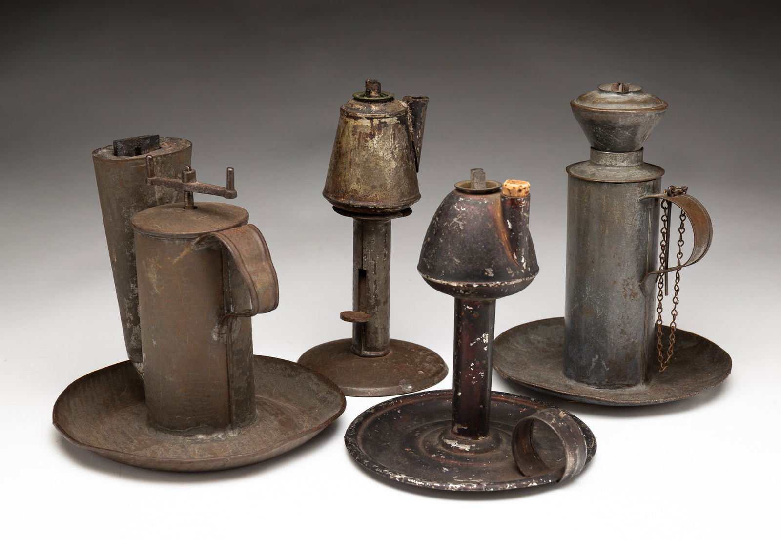 THREE WHALE OIL LAMPS AND A LARD LAMP.: American, 18th/19th Century. Three tin whale oil lamps: one a peg lamp on a hogscraper candlestick, one of cylindrical form with handle and attached wick pick, and one a Japanned chamber lamp with cor
