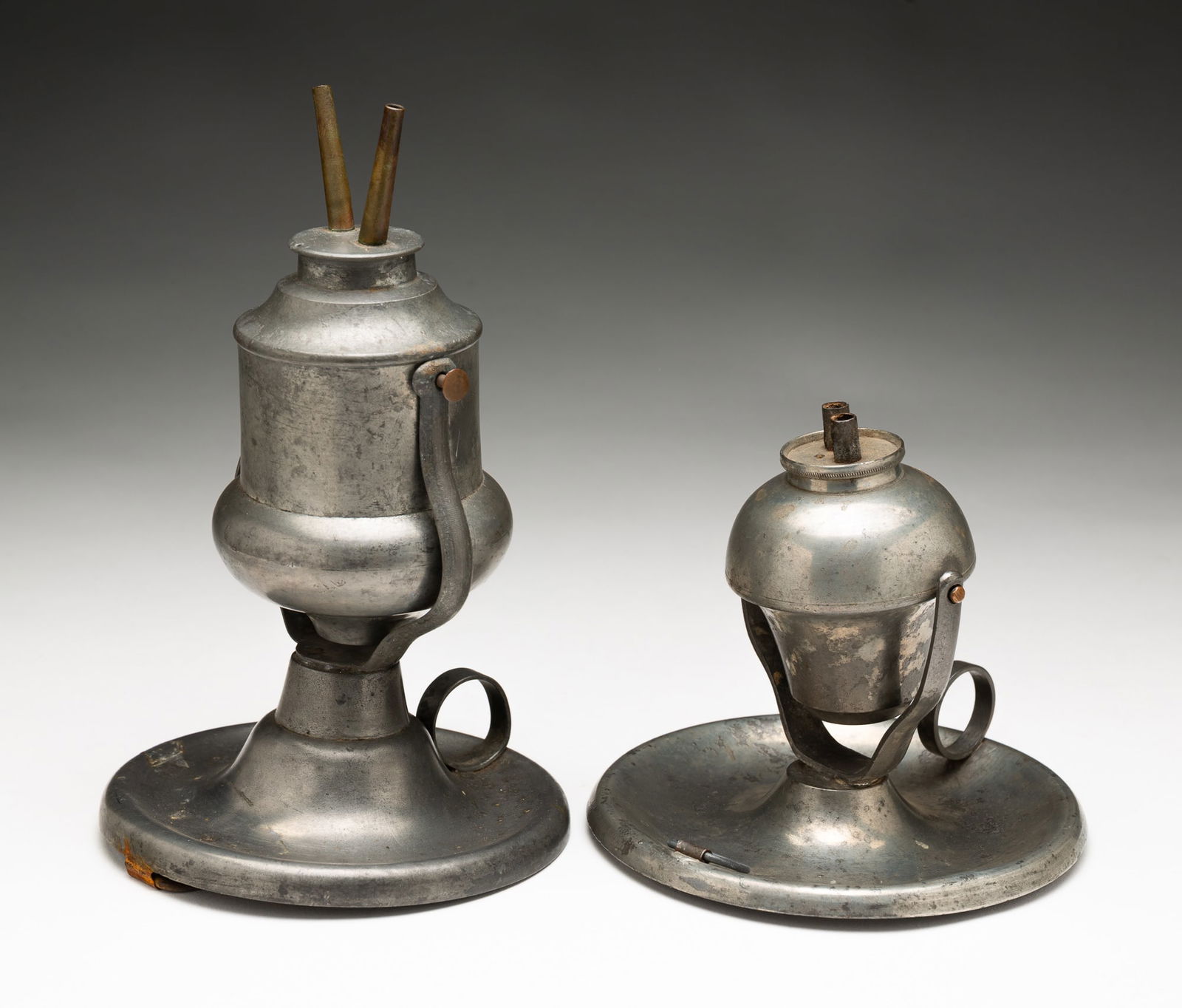 TWO GIMBAL MOUNT PEWTER LAMPS.: American, 19th Century. Both have handled saucer bases and loops for hanging. One has camphene burners and acorn shaped font and is marked M. Hyde. 7.75"h. And the second has double whale oil burner a