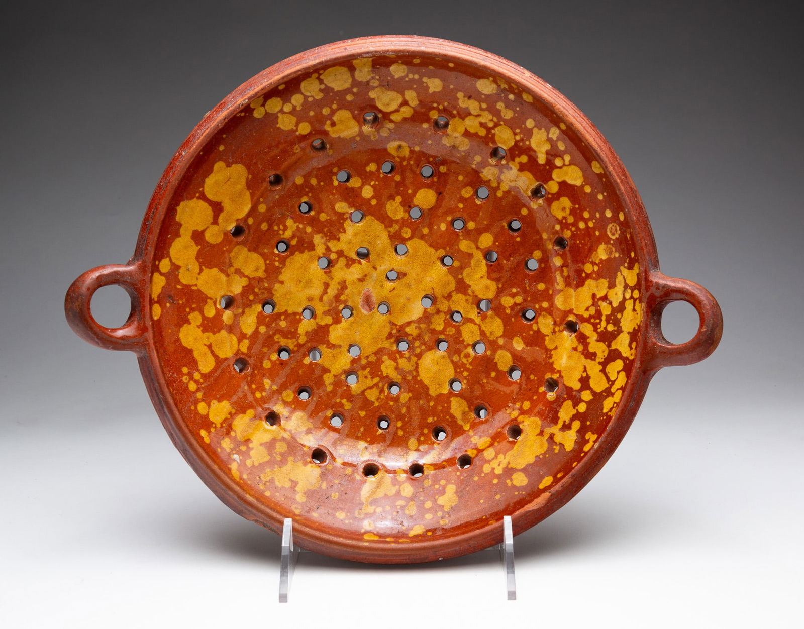 REDWARE COLANDER WITH SLIP DECORATION.: American, 19th Century. Two-handled shallow colander with banded rim, glazed in orange with yellow slip splatters. 3"h. 14.25"w.