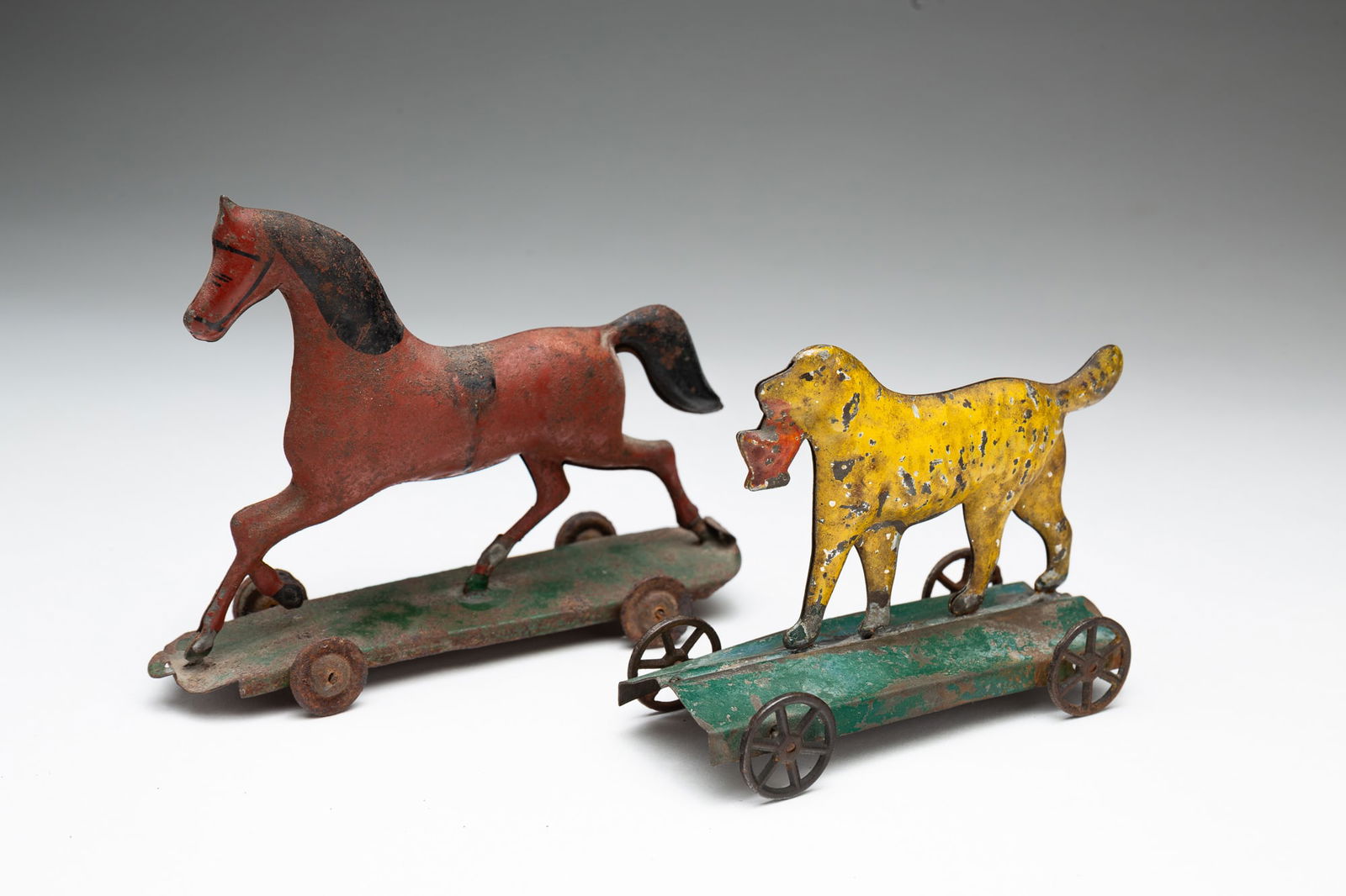 TWO PRESSED TIN AND PAINTED PULL TOYS.: American, late 19th Century. A yellow dog carrying an orange colored basket on a green base with spoked wheels. 3.25''h. 1.75''w. 4.25"l. And brown horse with black tail on a green base with solid whe