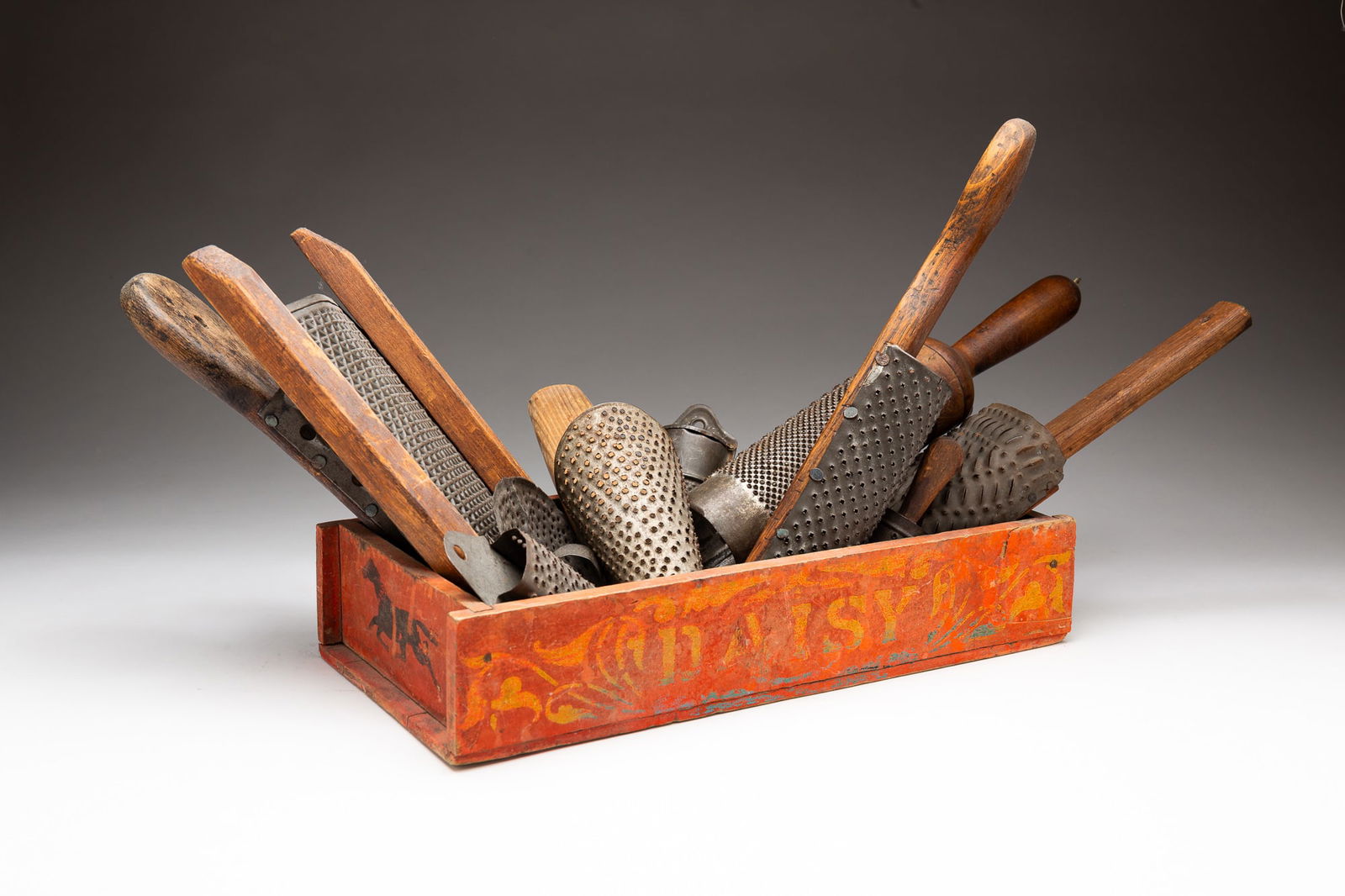 DAISY WAGON BED AND A COLLECTION OF GRATERS.: American, 19th Century. Rust red painted wagon bed with yellow flourishes and "Daisy" on sides, horses on ends and green interior. 2.75"h. 14"w. 7"d. Together with a collection of tin and wood and tin