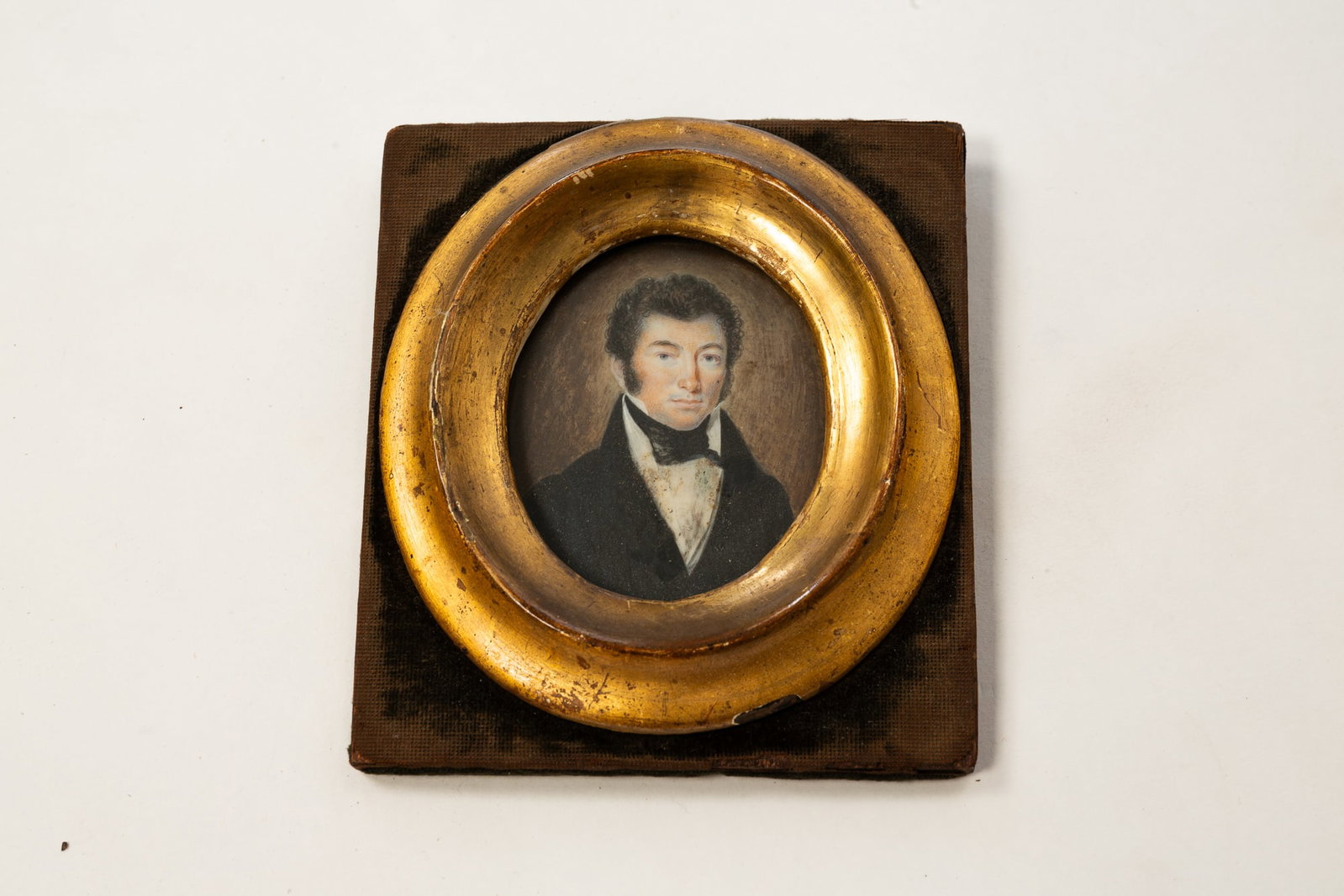 MINIATURE WATERCOLOR PORTRAIT OF A GENTLEMAN.: France, 19th Century. A gentleman with curly hair and bushy sideburns wearing a white shirt, black cravat, and black coat; on grayish brown background. In an oval gilt wood frame on a rectangular blac