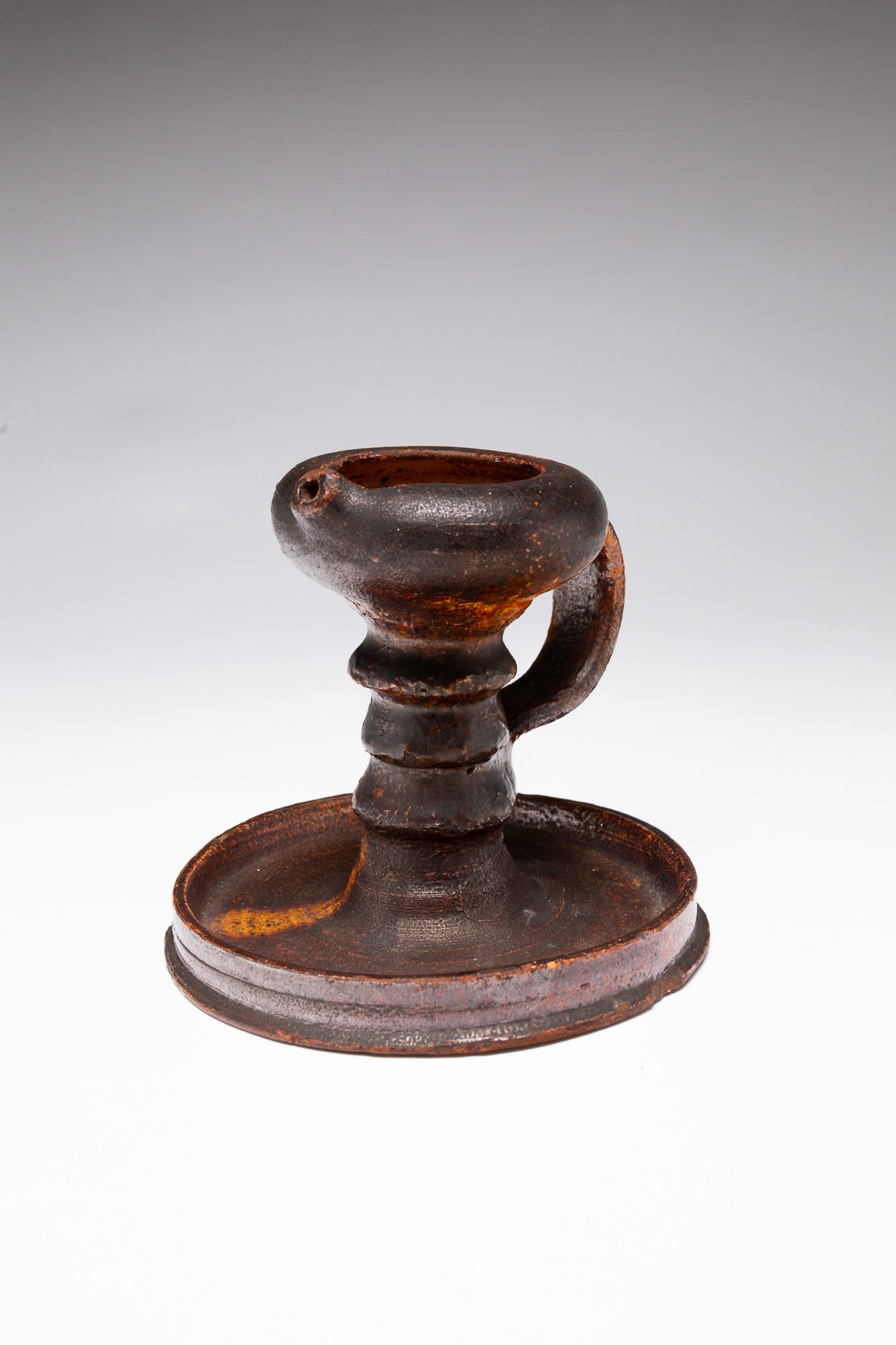 REDWARE BETTY LAMP.: American, 18th Century. Round form with applied handle, banded stem, and stepped base with an all over mottled yellow glaze. 5.25"h. From The Lifetime Collection of Mike & Marti Korba (Russiaville, In