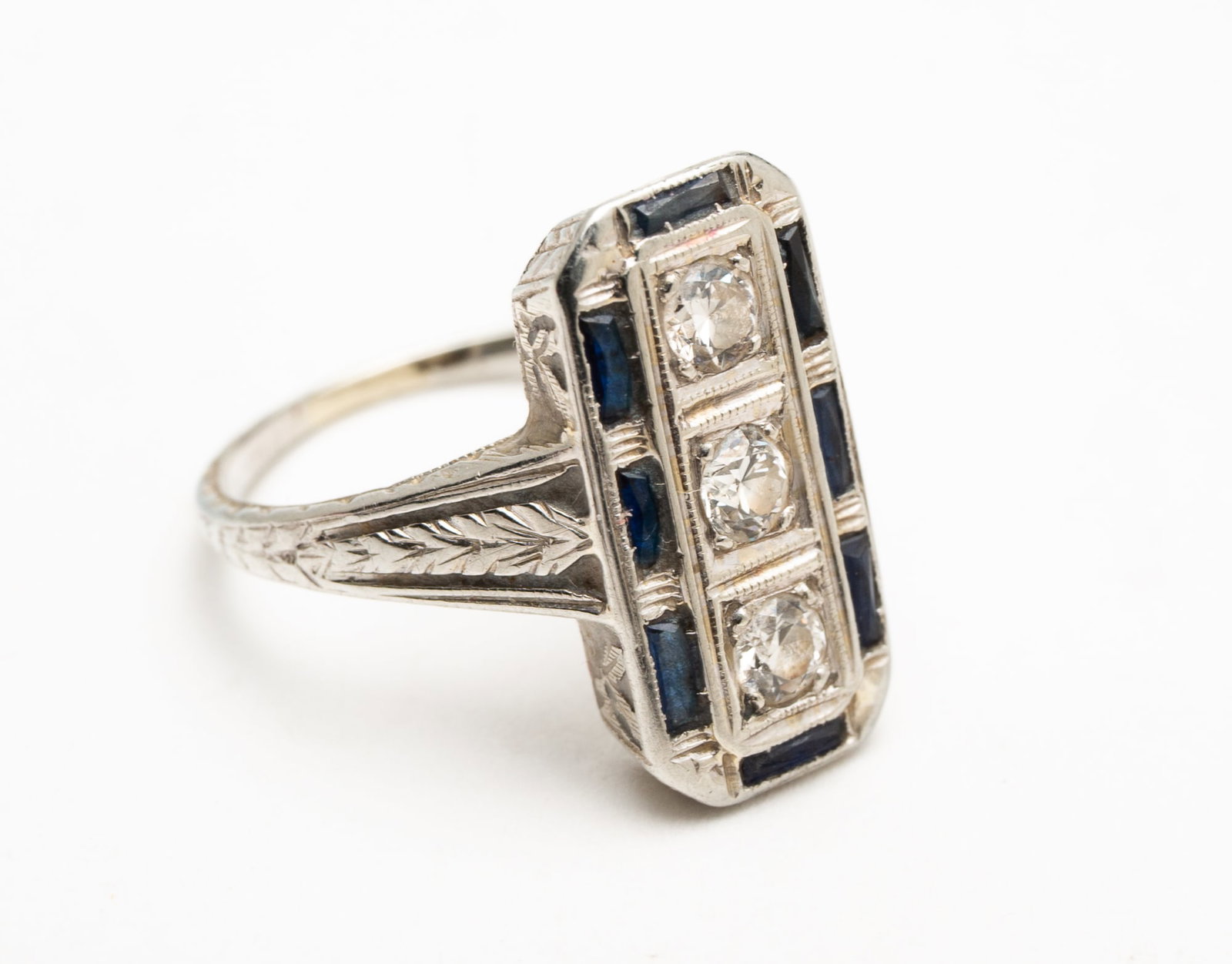 ART DECO WHITE GOLD RING WITH DIAMONDS AND SAPPHIRES.: Early 20th century. Stamped '19K' white gold, rectangular shaped ring. Three round cut diamonds down center, each approx. .10ct, border with seven faceted sapphire baguettes. Mounting with nice etchin
