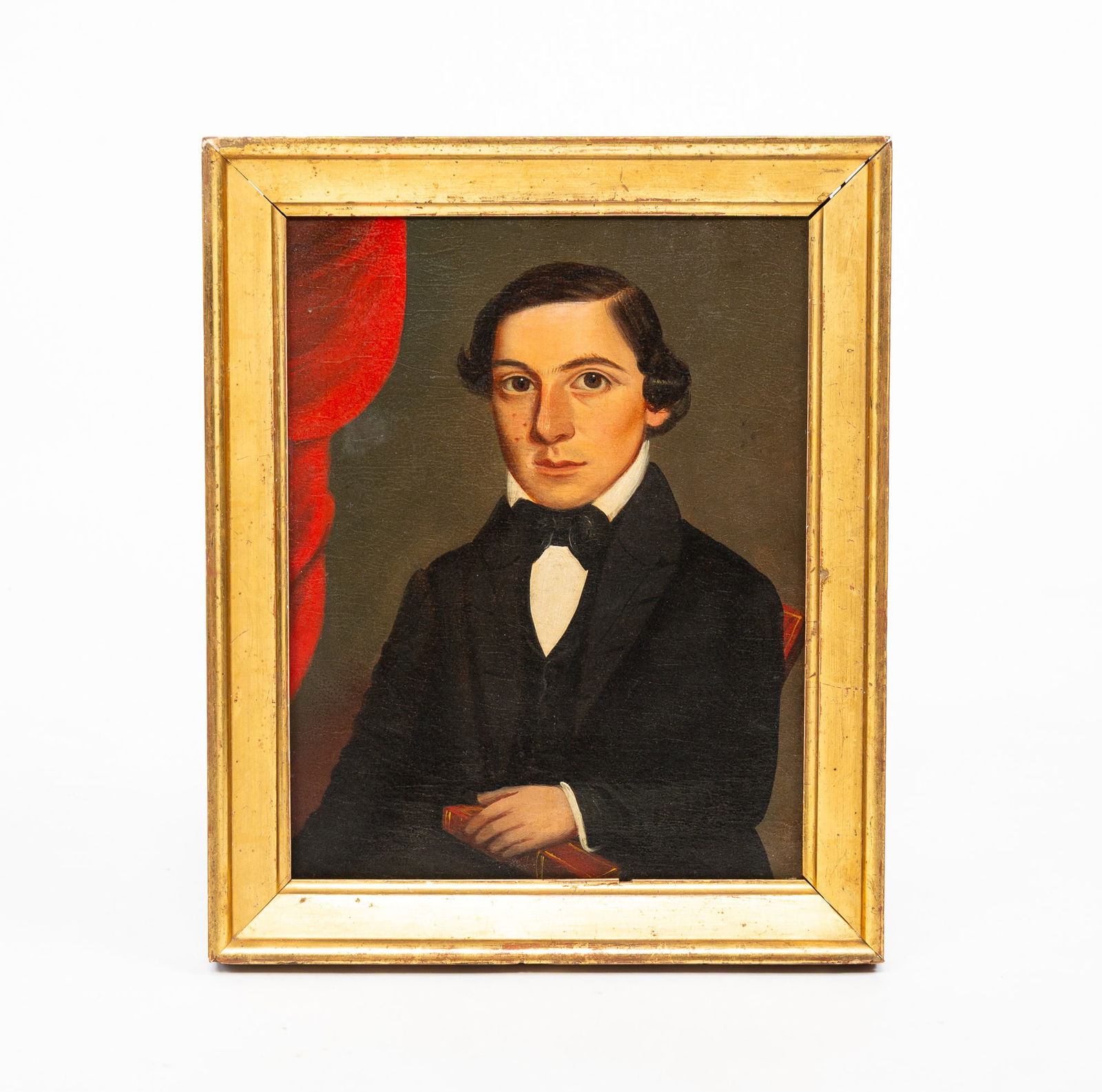 AMERICAN SCHOOL OIL ON CANVAS PORTRAIT OF A YOUNG MAN.: American, Mid-19th Century. Oil on canvas. A seated figure with curly dark hair and brown eyes wearing a black suit and bow tie. He holds a book and is seated before a red-draped background. 13.75"h.