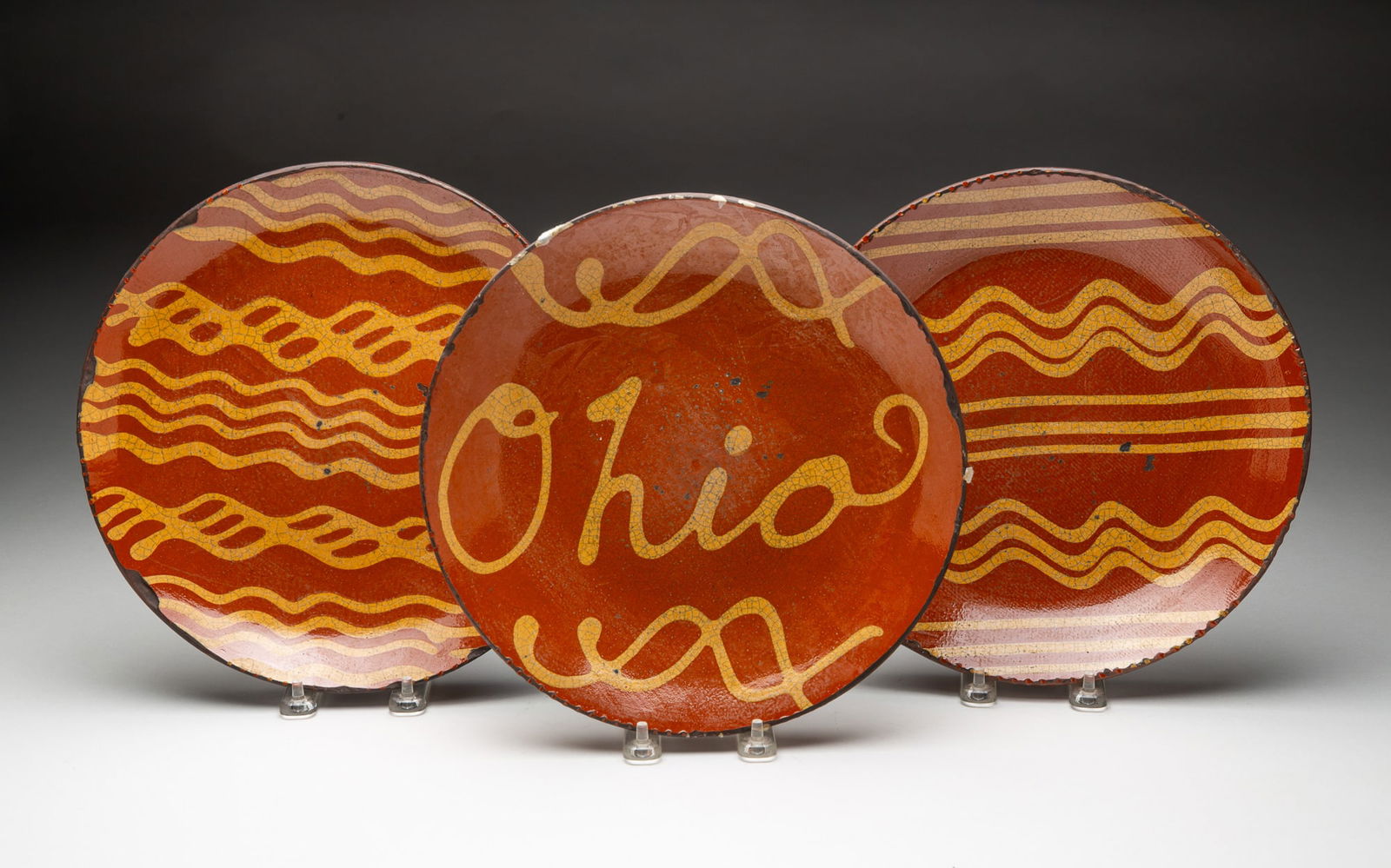 THREE GREG SHOONER SLIP-DECORATED REDWARE PLATES.: American (Ohio), Late 20th Century. Round plates in orange and black mottled glaze, two decorated with rows of yellow squiggly lines and one with the word "Ohio," all with coggled rims, all signed and