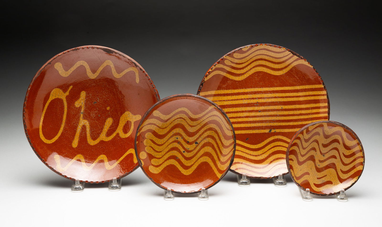 FOUR GREG SHOONER SLIP-DECORATED REDWARE DISHES.: American (Ohio), Late 20th Century. Three round plates in mottled orange glaze decorated with rows of yellow squiggly lines and one bowl in mottled orange glaze with the word "Ohio," all with coggled