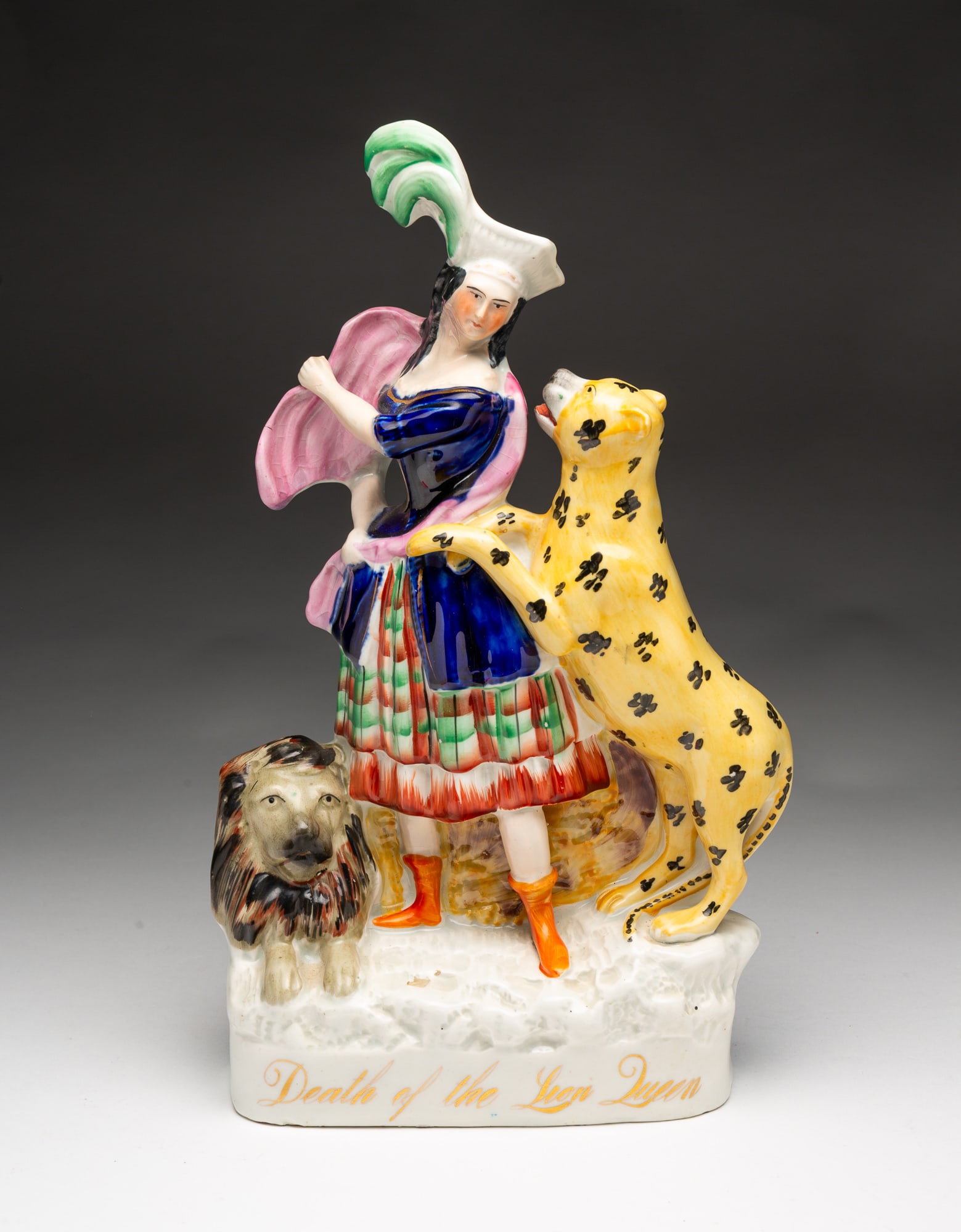 STAFFORDSHIRE "DEATH OF THE LION QUEEN" FIGURINE.: England, 19th Century. Lady in a Tartan plaid skirt, blue jacket and purple cape and plumed hat, a lion at her feet and a lioness in attack position on her left. 14"h.