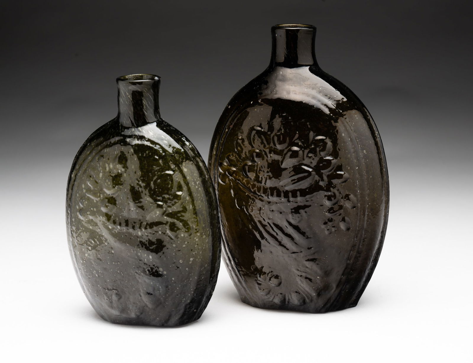 TWO AMERICAN OLIVE GREEN FLASKS.: American, 19th Century. One is eagle and cornucopia with broken pontil, 4.5"h. And a basket of flowers and fruit flask, broken pontil, chip on lip and not a clear impression, 5.75"h.