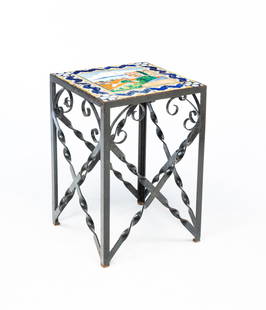 SPANISH COLONIAL STYLE WROUGHT IRON AND TILE TOP GARDEN TABLE.: 20th Century. Black-painted wrought iron table topped with glazed tiles depicting a seaside town with a mountain in the distance. 25"h. 17"w. 17"d. From the collection of Mark and Sandy Keily.
