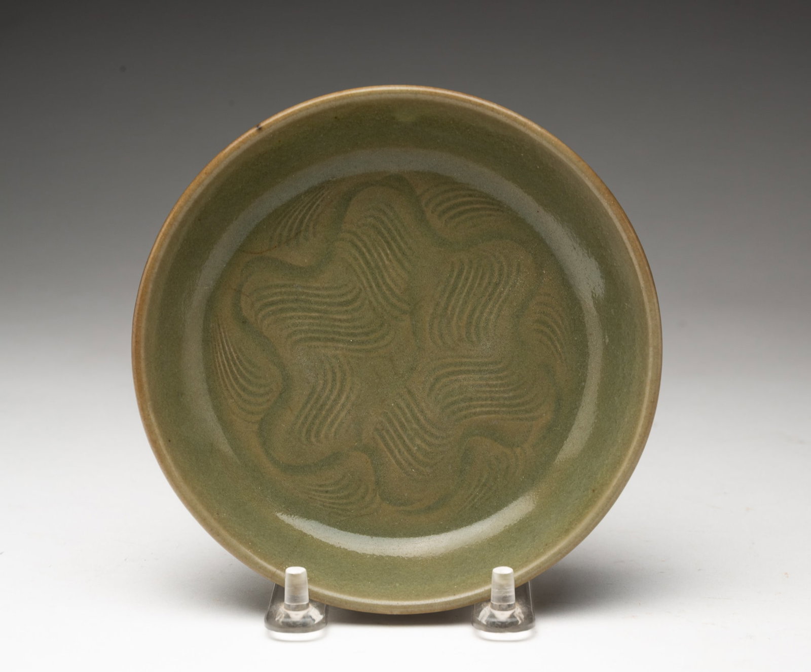 CHINESE YAOZHOU INCISED CELADON SAUCER.: China, probably Song Dynasty (10th to 13th Century). Of shallow, circular form with incised wavy lines in a pinwheel-type pattern on the interior, covered in a green glaze, the bottom with an inverted