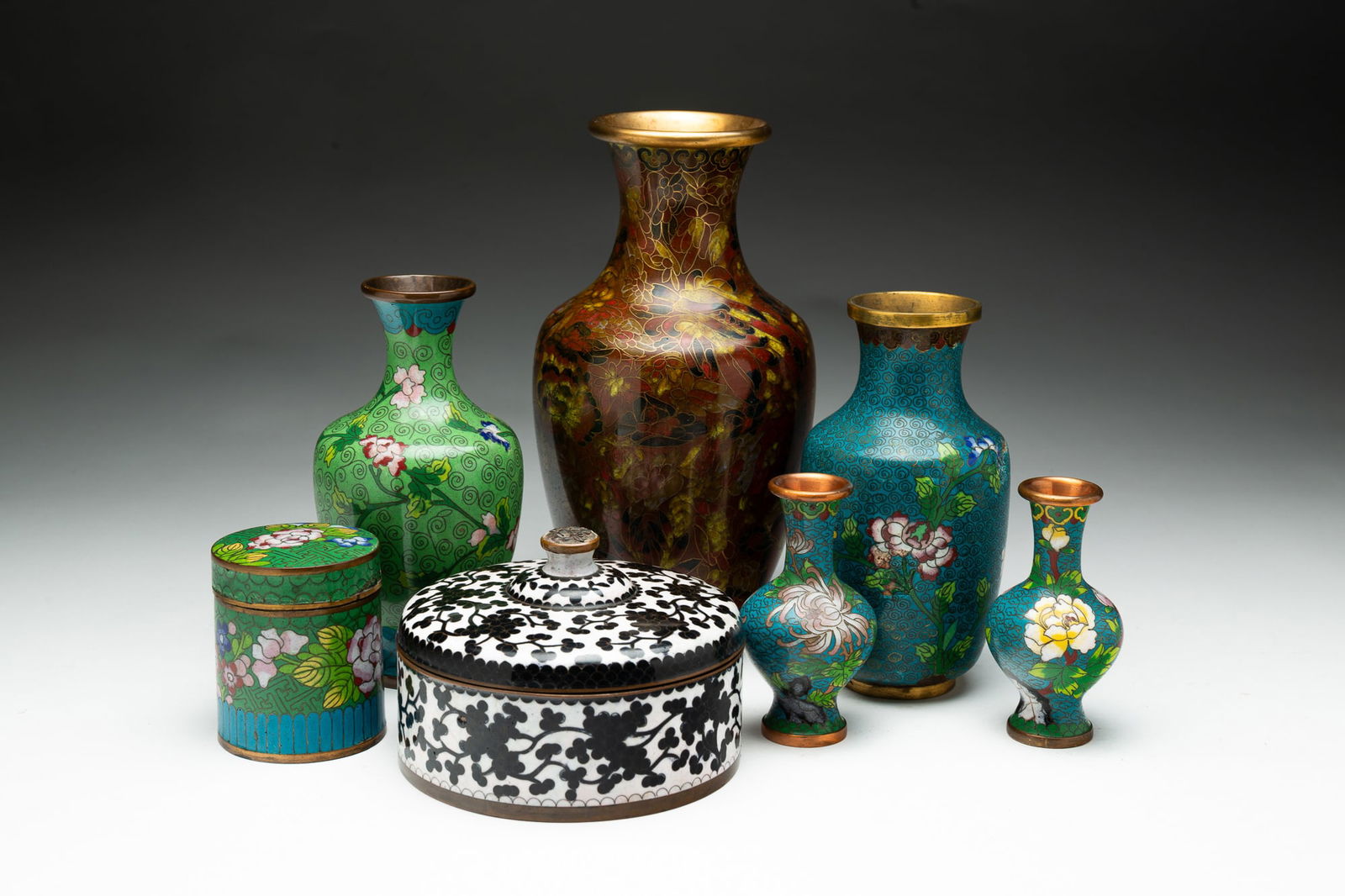 SEVEN CLOISONNE ITEMS.: China, 20th Century. All seven have floral enamel decorations. Three vases, 9.25"h., 6.75"h. and 6.5"h. A pair of bud vases, 4"h. Cylindrical jar, 3"h. And a black and white jar with knob finial, 3.25