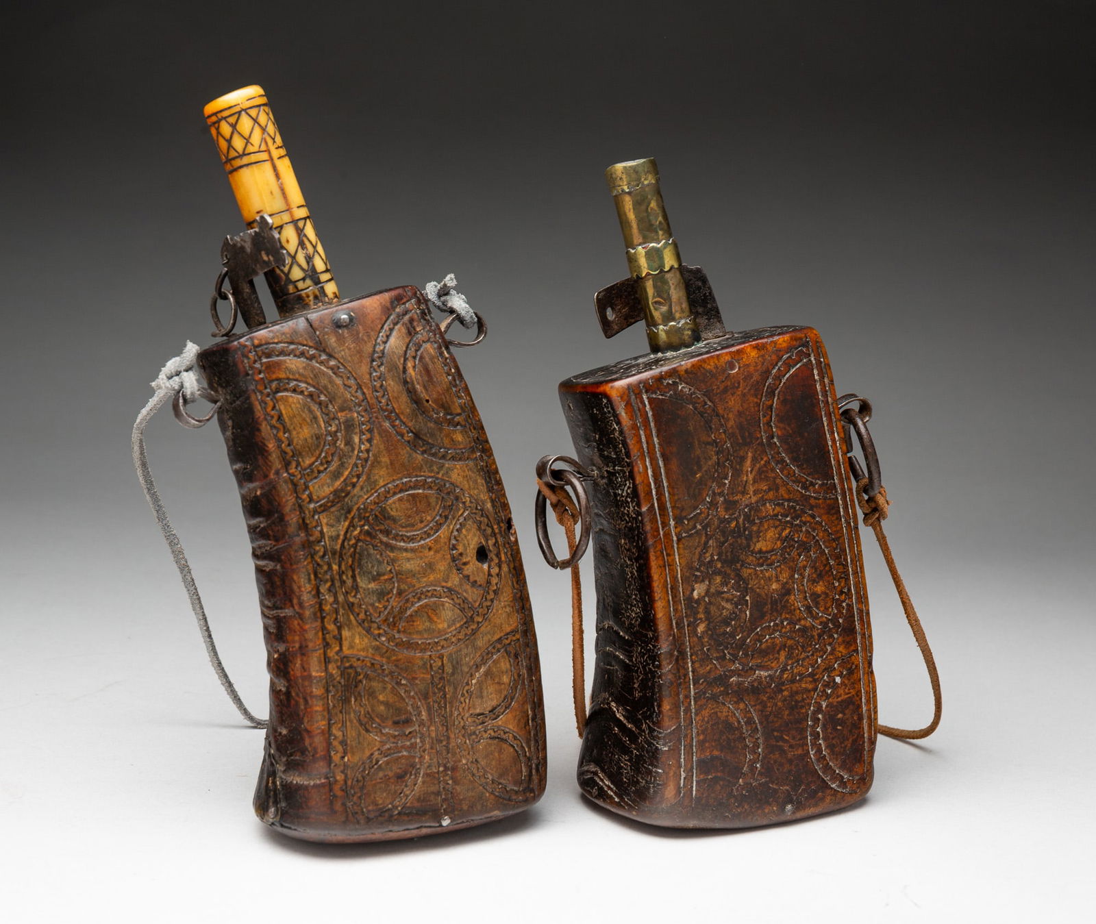 TWO NORTH AFRICAN HORN POWDER FLASKS.: North Africa, ca.1900. Of irregular rectangular form having carved circles and arcs, with iron fittings and leather straps, one having a bone spout and the other brass. The larger, 9"h.