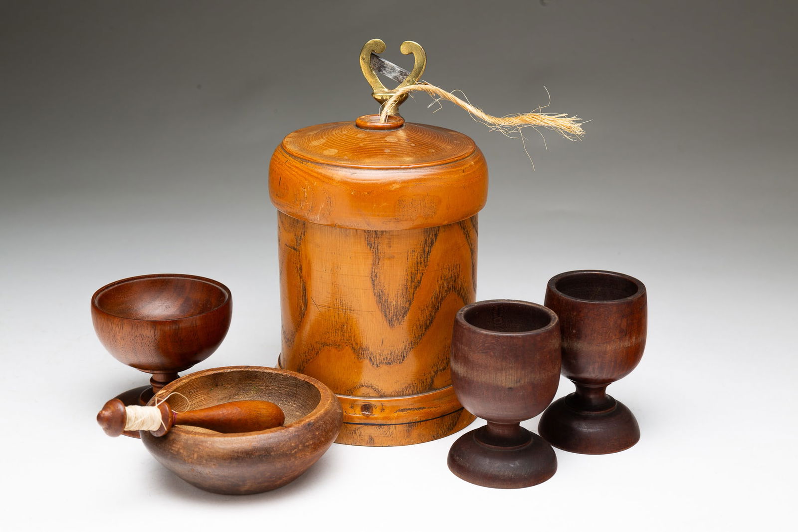 SIX AMERICAN TREEN ITEMS.: American, 19th and 20th Centuries. Cylindrical string holder with heart shape cutter. 7"h. Two footed egg cups, 3"h. A miniature bowl, 1.5"h. 3.75"I. Footed jar, 2.5"h. And a bobbin, 4"h.