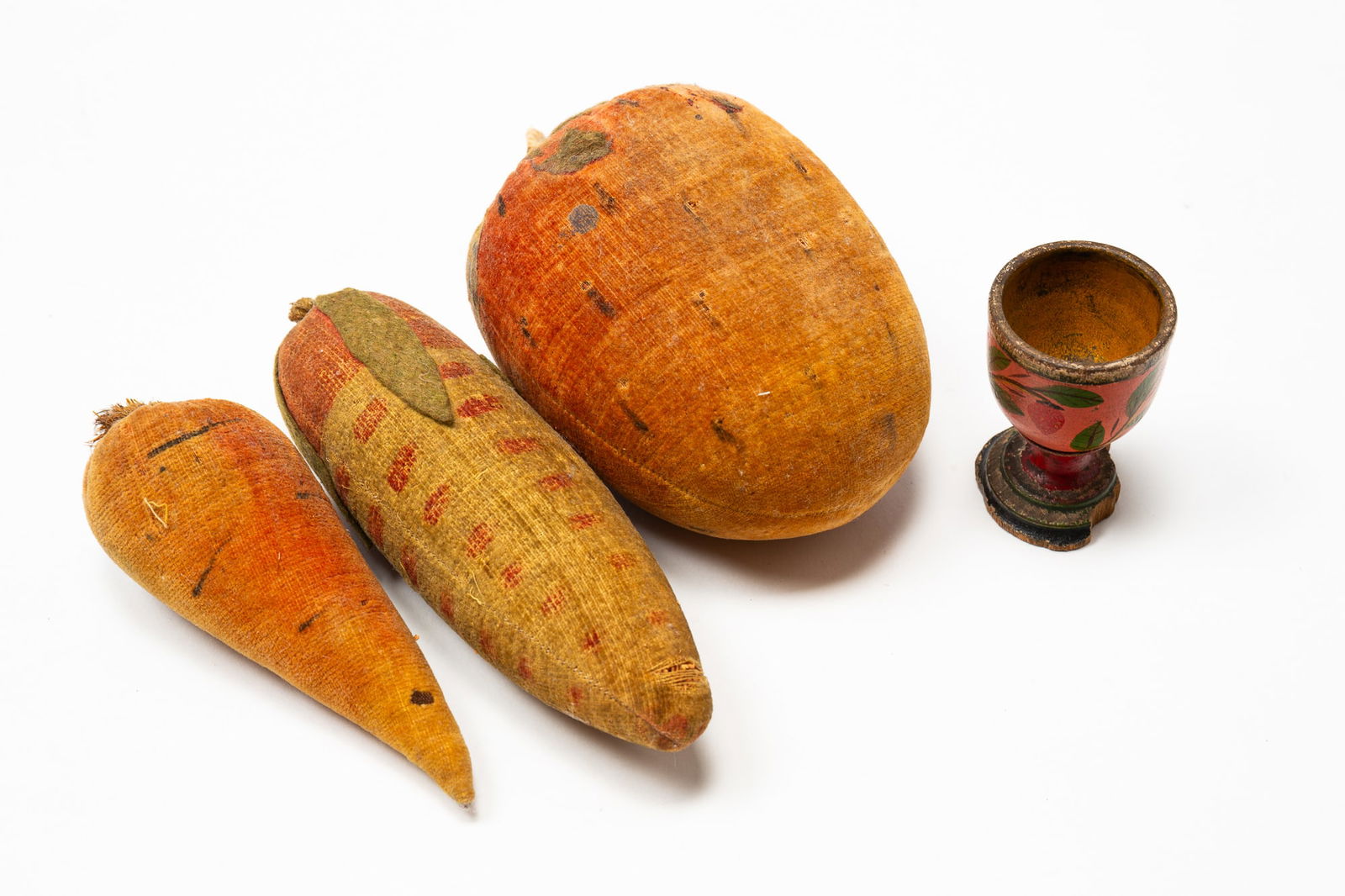 AMERICAN LEHNWARE EGG CUP AND THREE PINCUSHIONS.: American, 19th Century. Egg cup with painted strawberries on a pink background, yellow interior and stepped foot (damage to foot). 2.5”h. Three pincushions: ear of corn, 6.25"l; carrot, 5”