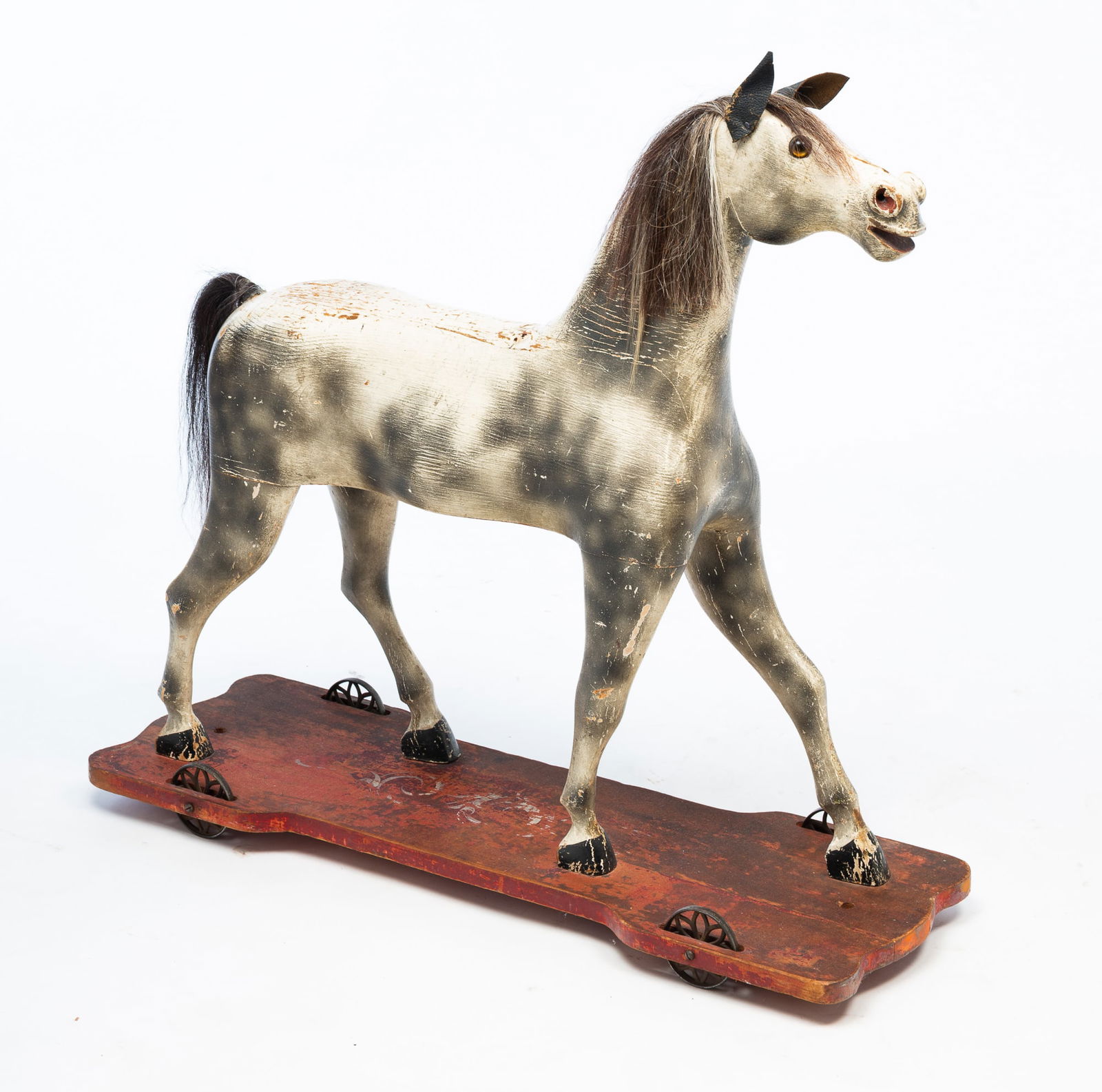 LARGE GERMAN CARVED AND PAINTED PINE HORSE PULL TOY.: German, Late 19th Century. Pine. Multi-section horse with white and gray dappling, horsehair mane and tail, glass eyes, and leather ears. Shaped base with worn red paint and white flourishes with four