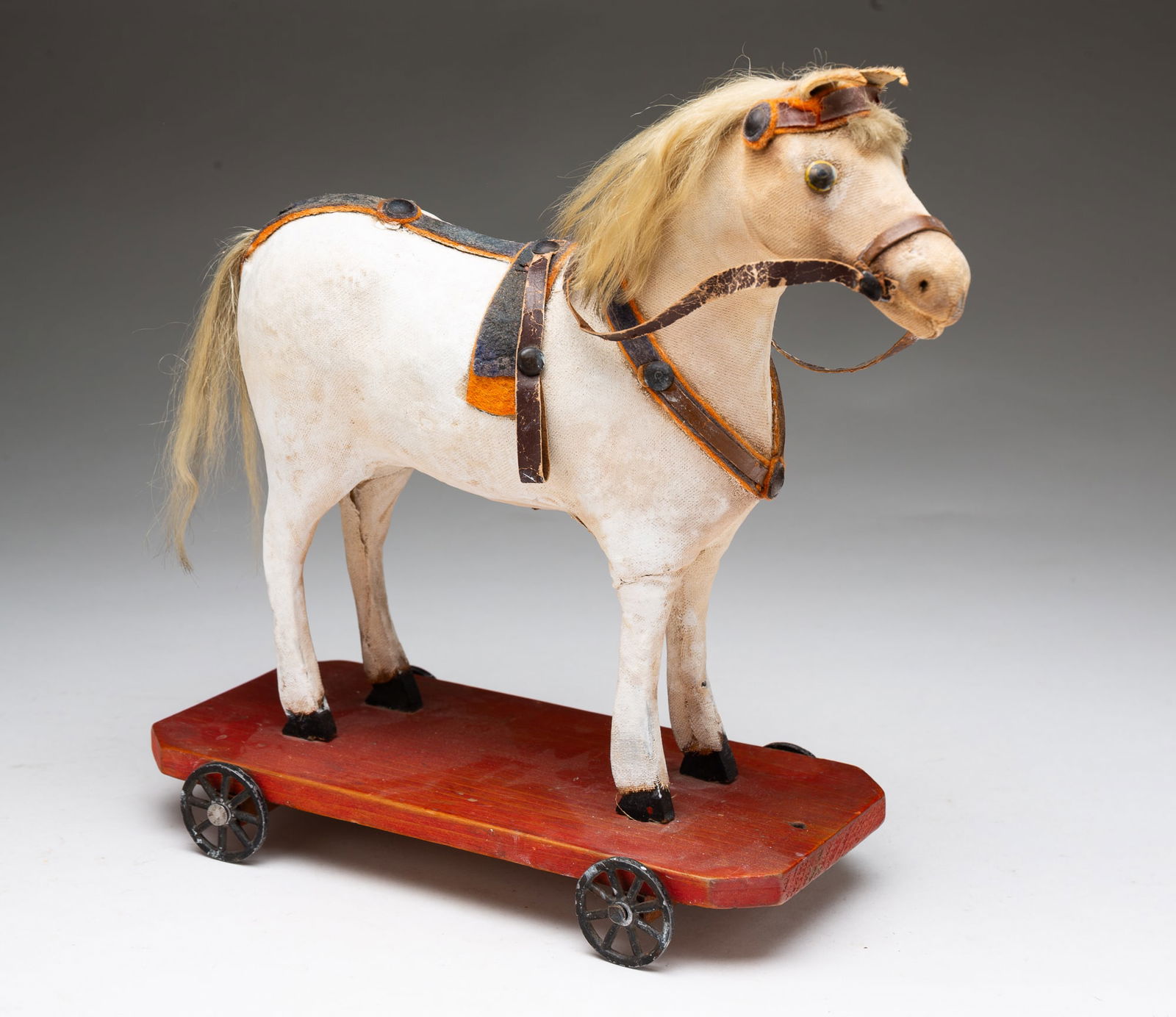 GERMAN CLOTH-COVERED PAPIER-MACHE HORSE PULL TOY.: Germany, Early 20th Century. Having a papier-mache body covered in white-painted cloth, the legs of wood, with metal eyes, leather ears, leather reins, an synthetic mane and tail, mounted on a red-was