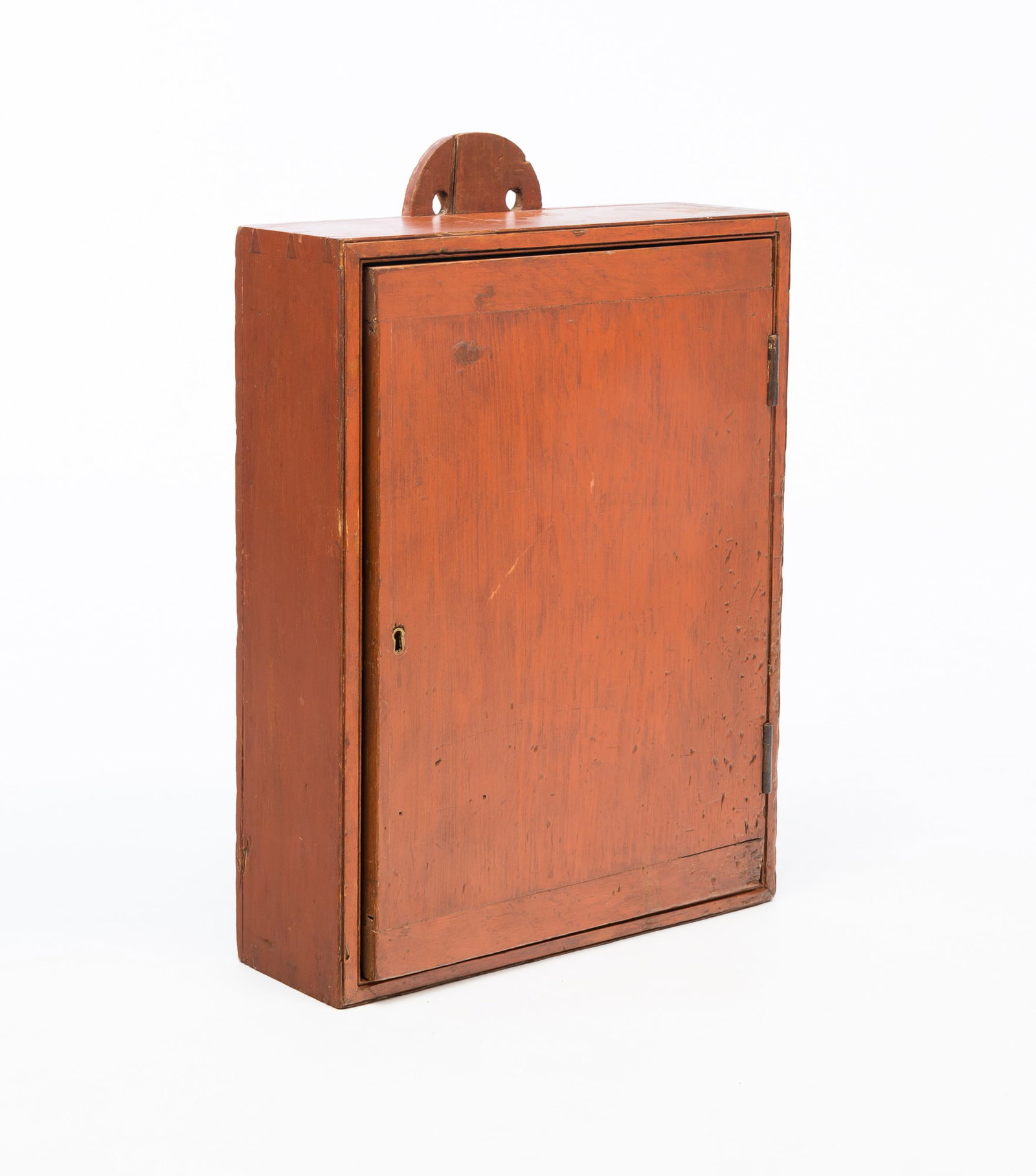 AMERICAN REDDISH-ORANGE PAINTED PINE HANGING CUPBOARD.: American, Late 19th Century. Pine. Dovetailed case, arched tab hanger with two holes, single one panel door with breadboard ends and keyhole, and original reddish-orange paint. 22"h. 17"w. 5.75"d.