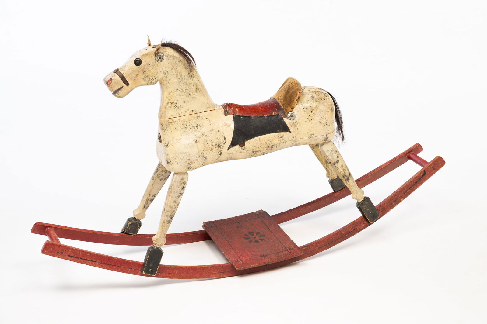 AMERICAN CARVED AND PAINTED WOOD ROCKING HORSE.: American, Early 20th Century. Pine. Multi-section horse with white dapple paint, horse hair tail, rivet eyes, leather ears, red leather saddle with velvet saddle seat and leather trim. Base with worn