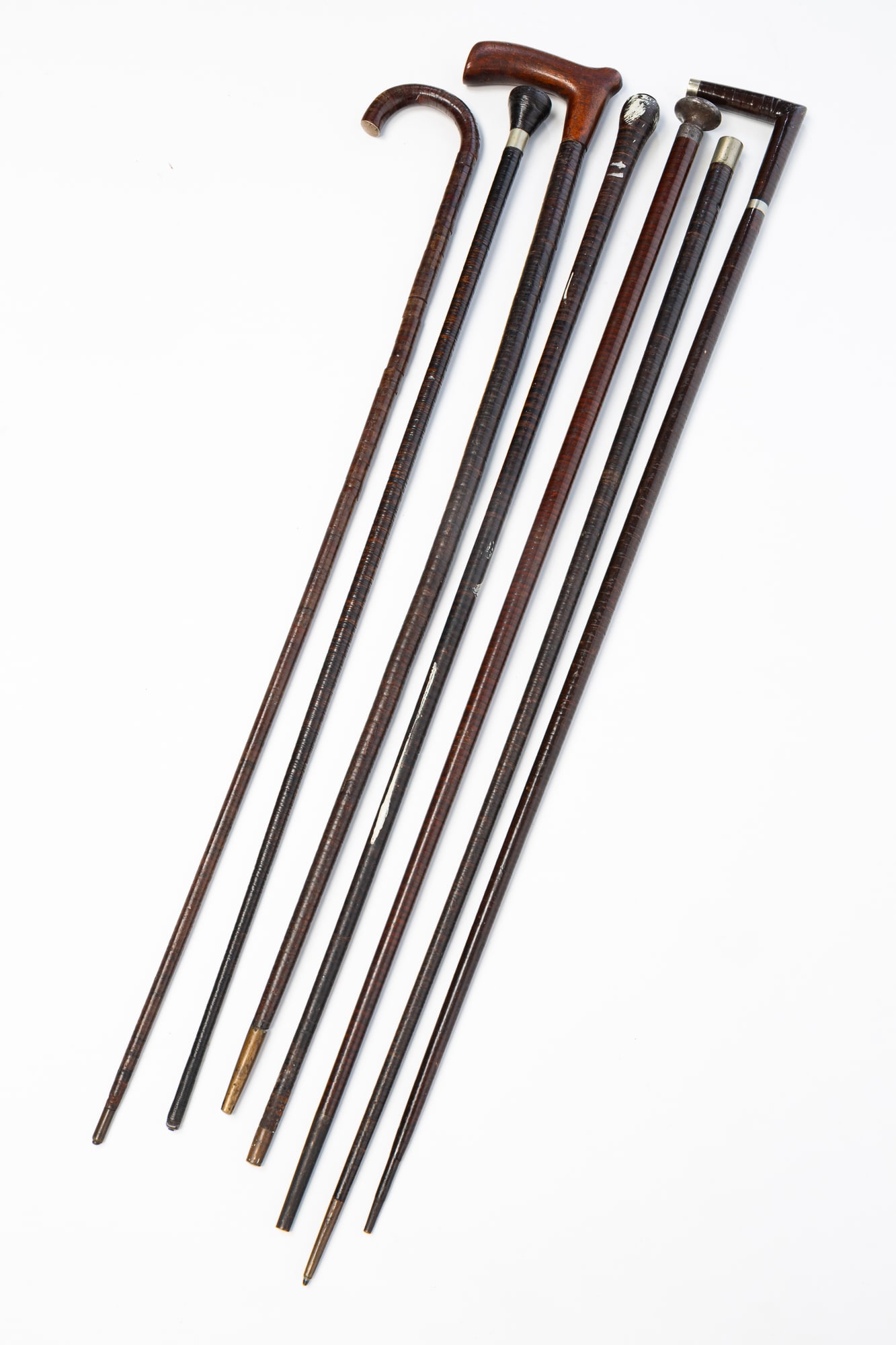 COLLECTION OF STACKED LEATHER CANES AND WALKING STICKS.: Late 19th-early 20th Century. Leather, wood, and metal. Comprising three handled canes and four walking sticks, three of which have a metal knob or collar. Some with brass ferules. Example with mahoga