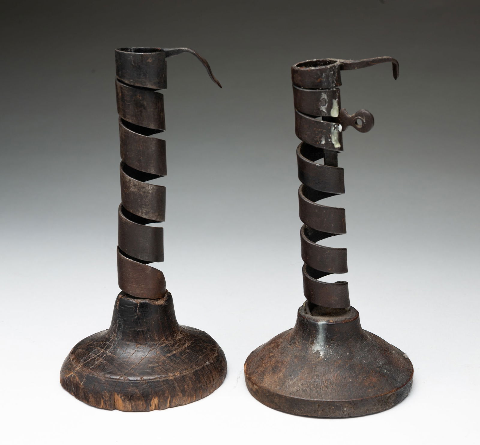 TWO AMERICAN WROUGHT IRON CANDLESTICKS WITH TURNED WOOD BASES.: American, ca. 1800. Wrought spiral candlesticks mounted on round wooden bases, the tops with hooks, one is adjustable. 7.5"h. and 7.25"h.Provenance: From the Lifetime Collection of the late Joanne & D