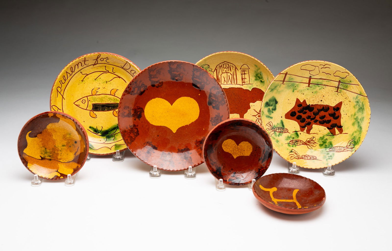SEVEN LESTER BREININGER JR. (ROBESONIA, PENNSYLVANIA, 1935-2011) SLIP-DECORATED REDWARE DISHES.: Lester Breininger Jr. (Robesonia, Pennsylvania, 1935-2011), 20th Century. Four plates all having coggled rims, an orange and black mottled glaze, and yellow slip decoration: a small bowl with an "H;"