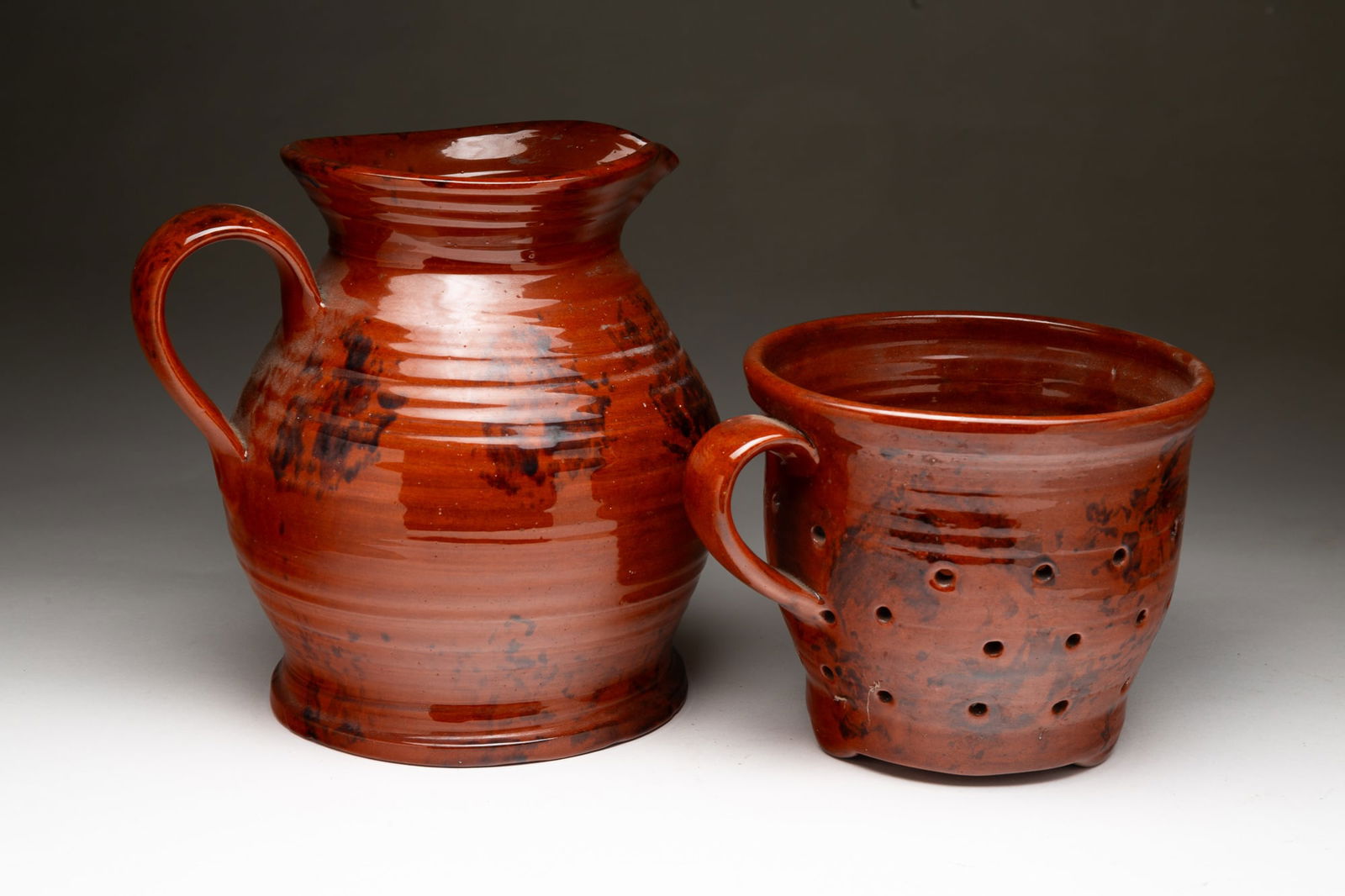 LESTER BREININGER JR. (ROBESONIA, PENNSYLVANIA, 1935-2011) REDWARE PITCHER AND COLANDER.: Lester Breininger Jr. (Robesonia, Pennsylvania, 1935-2011), 20th Century. Handled pitcher with an orange and black mottled glaze; signed and dated 1979 on underside. 9"h. 9.5"w. Handled colander with