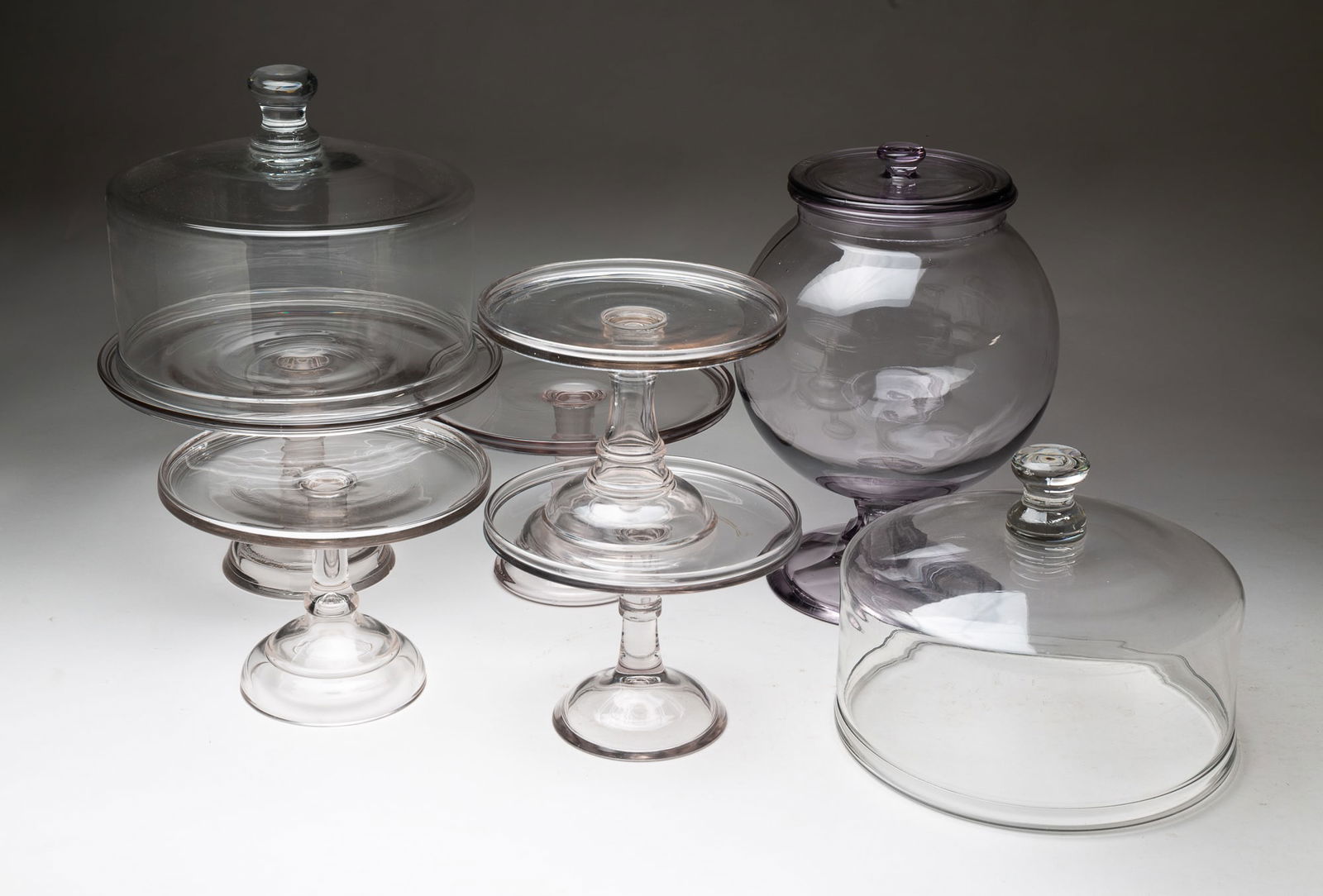 FIVE AMERICAN GLASS CAKE PLATES, TWO GLASS DOMES, AND A GLASS CANDY JAR.: American, ca. 1900. Five glass cake plates with pedestal bases ranging from 8.25"d. to 12.5"d. Two glass domes, both 11"d. And a purple glass candy jar with lid on a pedestal base. 14.5"h. 9"d.Provena