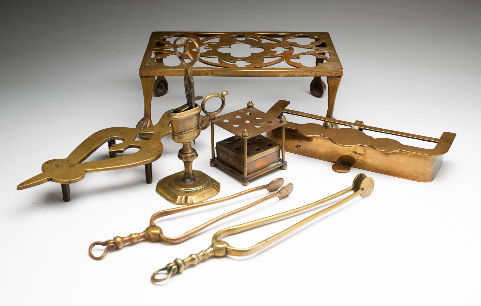 SEVEN BRASS HEARTH ITEMS, INCL. MINIATURE KETTLE STAND.: English and American, 19th Century. Comprising a miniature kettle stand, a miniature fire fender, kettle stand with claw feet, two pairs of tongs, a trivet, and a wick trimmer with stand. Largest (ket