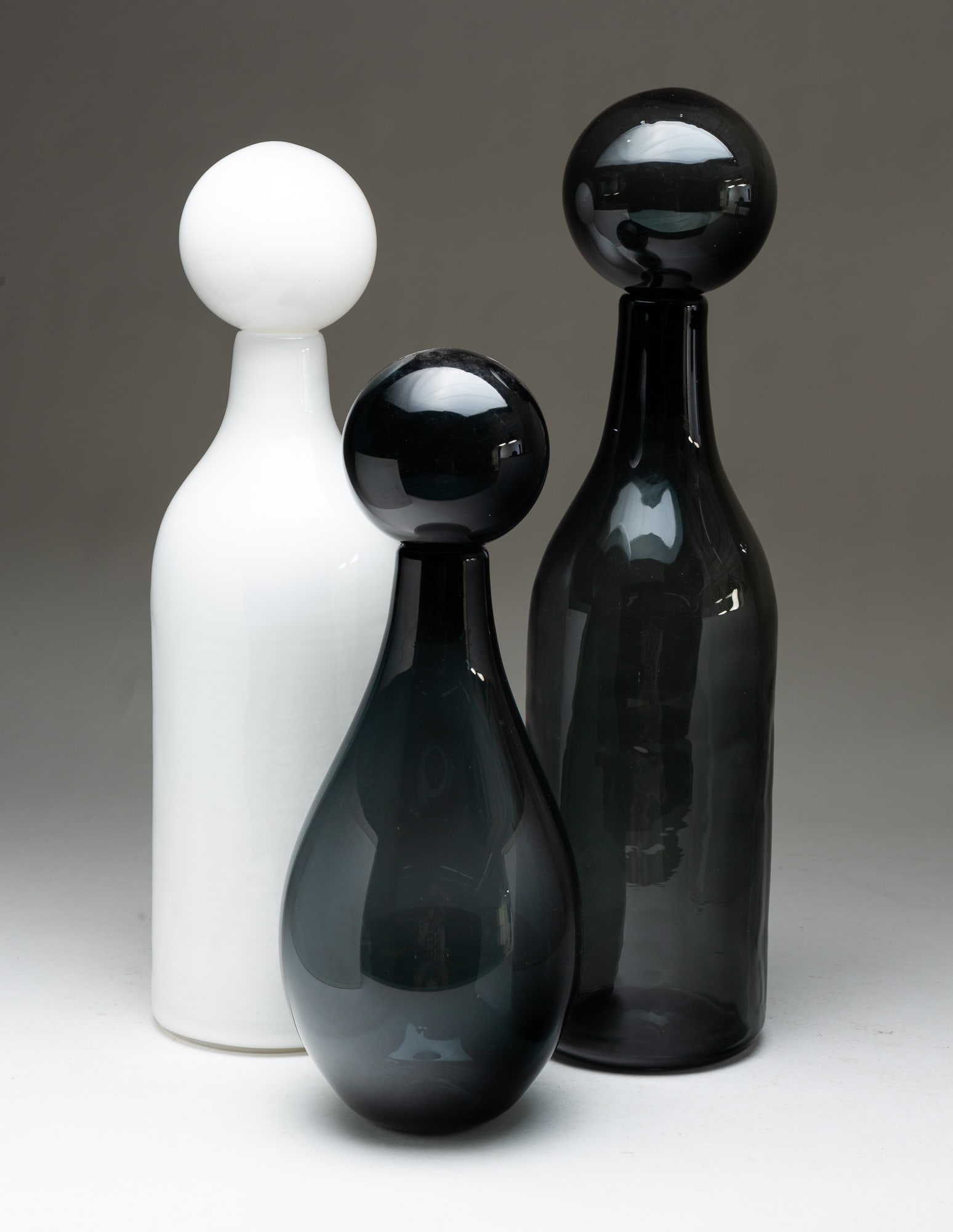 ELIZABETH LYONS (AMERICAN, 20TH/21ST CENTURY), SET OF THREE BLOWN GLASS OVERSIZED DECANTERS.: American (New York), contemporary. Blown glass. Two in smoky glass and one in white. Large glass bottles with spherical stoppers. Signed to bottoms. 19"h. to 14.5"h.