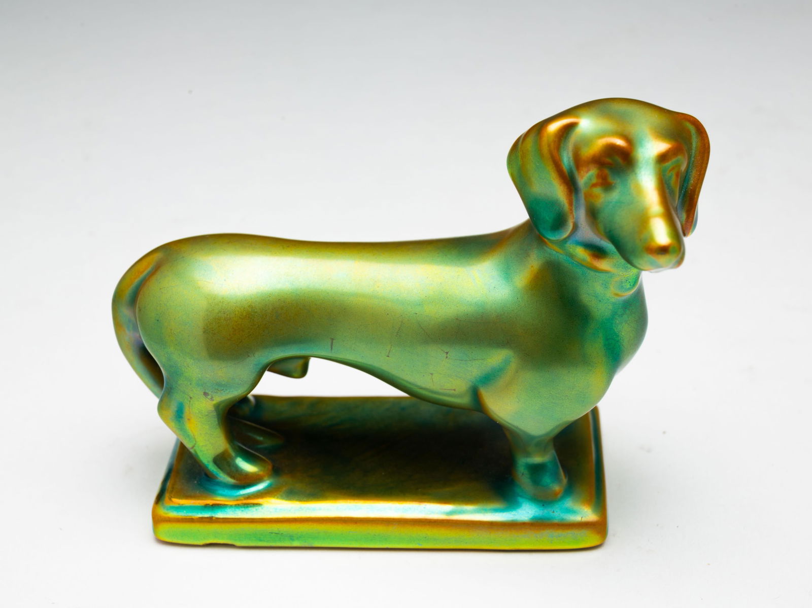 ZSOLNAY HUNGARIAN PORCELAIN DACHSUND FIGURINE.: Hungary, Early 20th Century. Dachsund figurine on rectangular base having an iridescent green glaze, signed on underside. 3"h. 4"w. 1.75"d.