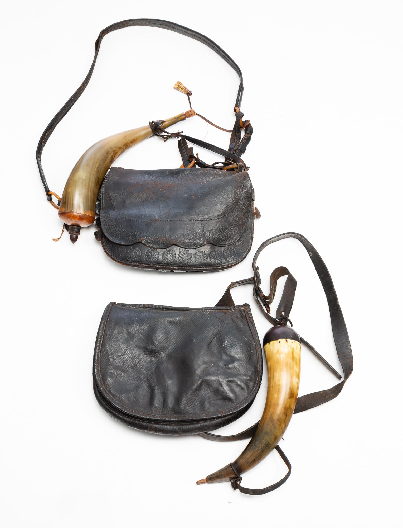 TWO LEATHER HUNTING BAGS WITH POWDER HORNS.: American, 19th Century. Leather, horn, and wood. Each bag with a shoulder strap continuing to a fold-over flap concealing a storage pocket. One has a tooled exterior with a carved antler shot measure