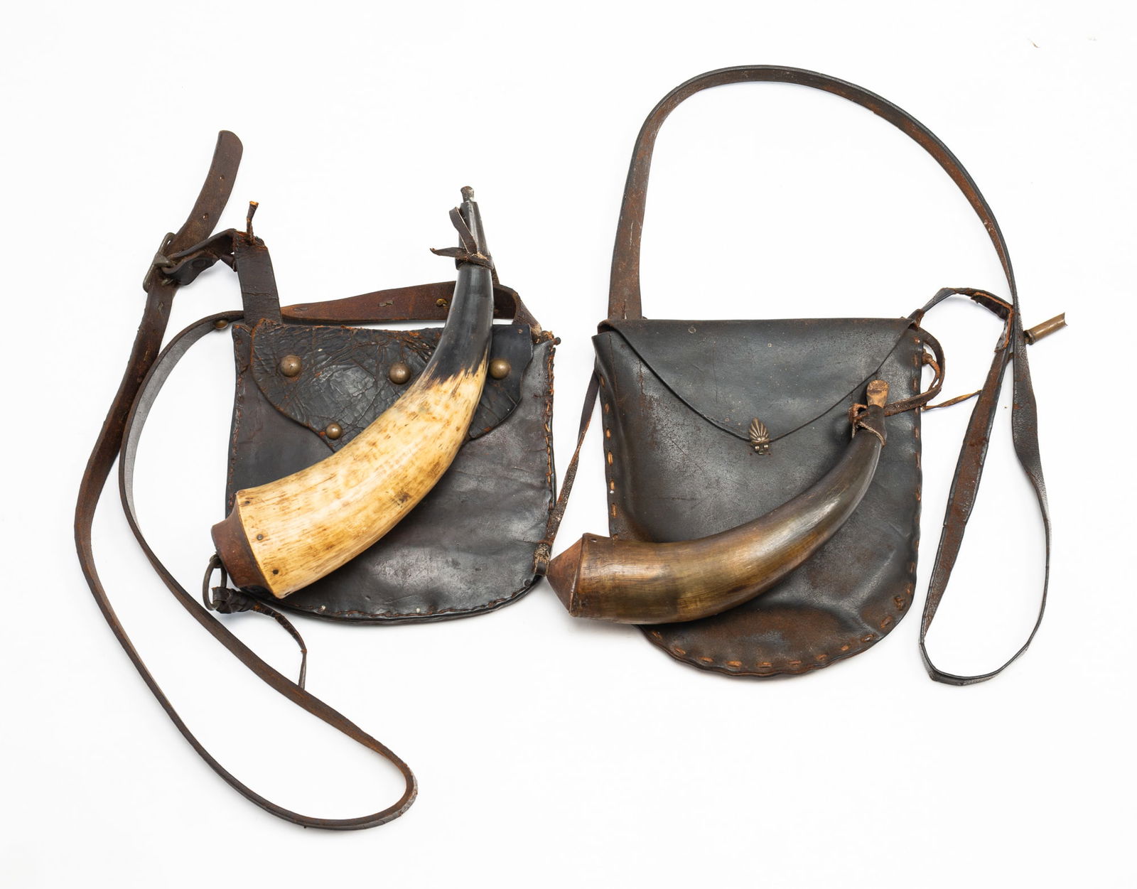 TWO LEATHER SINGLE-POCKET HUNTING BAGS WITH POWDER HORNS.: American, 19th Century. Leather, brass, horn, and wood. Each bag with a shoulder strap continuing to a fold-over flap concealing a single storage pocket. One has brass studs while the other has a bras