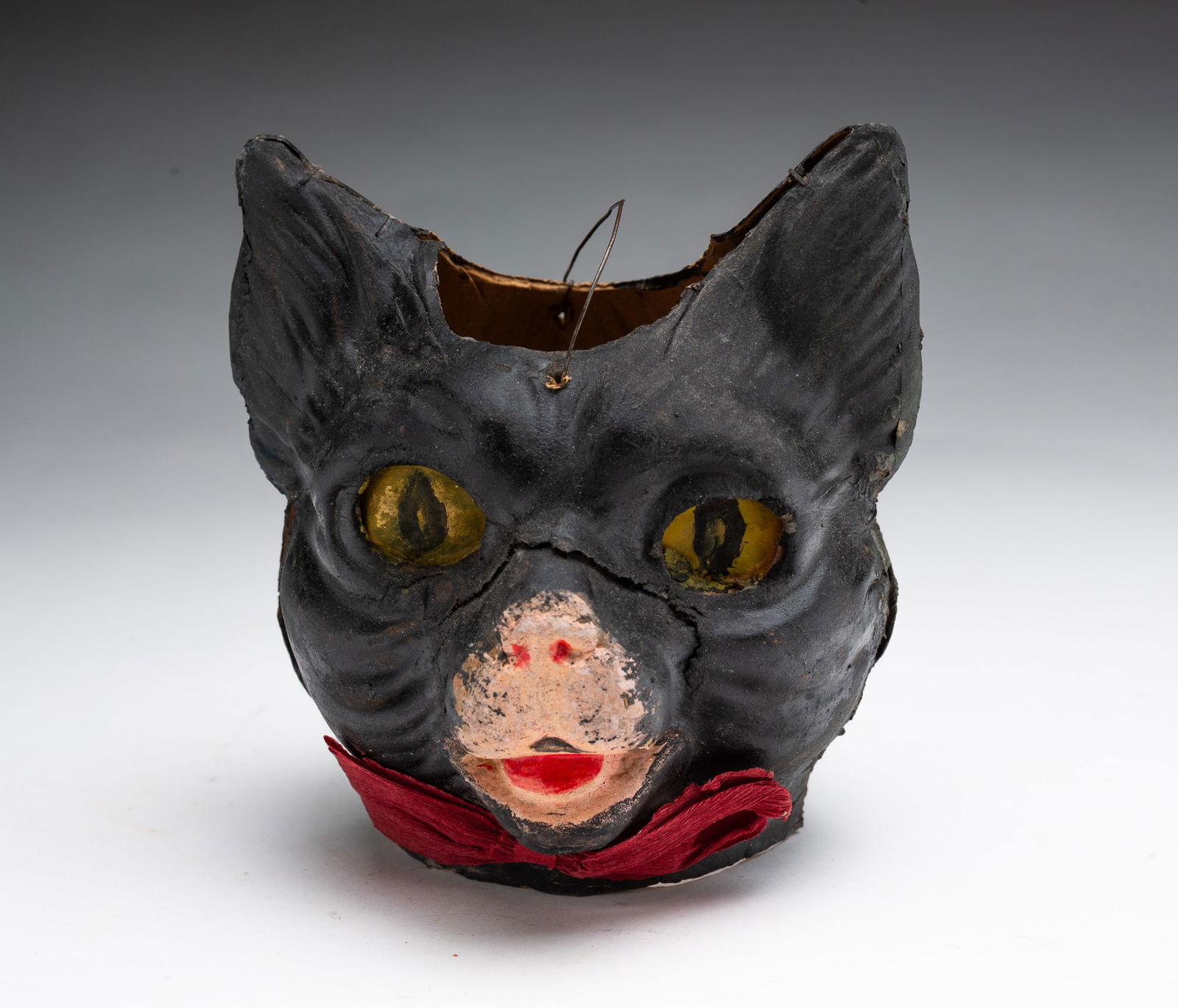 GERMAN PAINTED AND MOLDED PAPER BLACK CAT HALLOWEEN LANTERN.: Ca.1910. Molded paper in two parts held together with staples in the form of a black cat with red paper bow, pink nose, and yellow eyes, having a wire bale handle. 5.5"h. 5.5"w. 4.5"d.Provenance: From