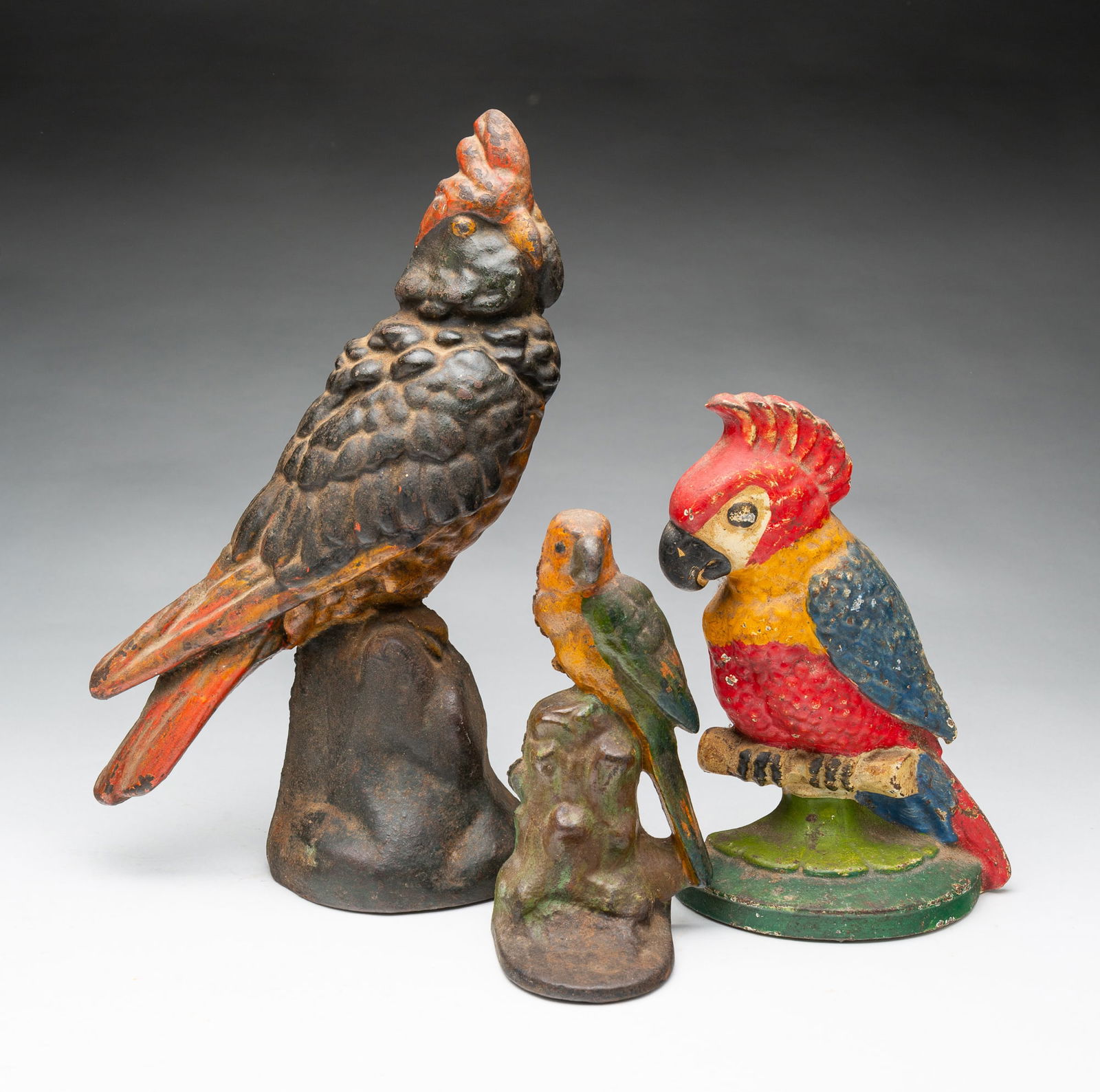THREE AMERICAN PAINTED CAST IRON PARROT DOORSTOPS.: Early 20th Century. Set of three parrots, the largest and smallest perched on a tree stump base, the middle on a perch with half-round base, all with original paint. Tallest, 12"h.Provenance: From the