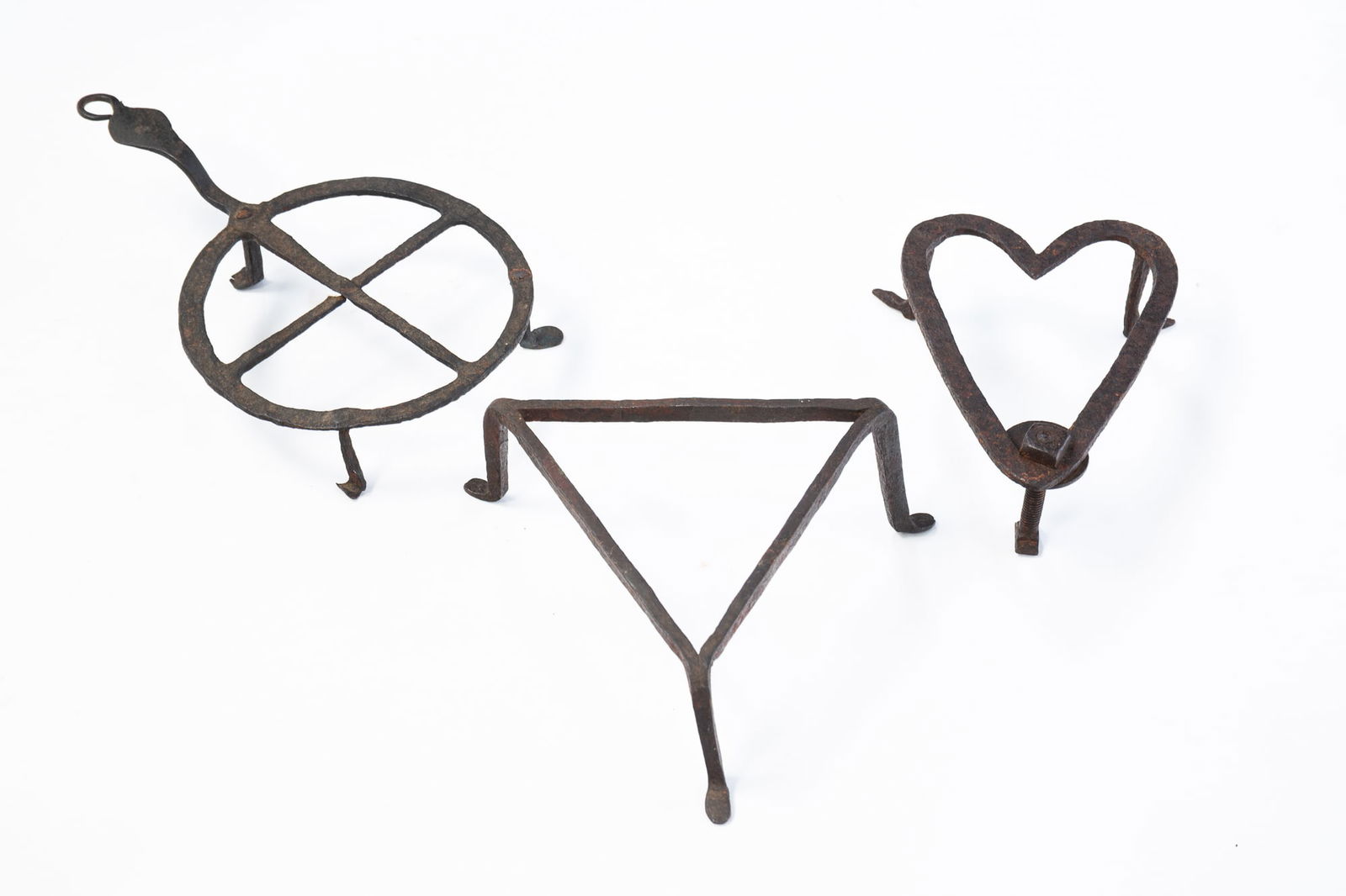 THREE AMERICAN WROUGHT IRON TRIVETS.: Mid-19th Century. In three forms; a heart with three legs including a make-do repaired leg, 2"h. 6"h. A circle with cross-hairs, 1.75"h. 9"l. And a tringle with three legs with shoe feet, 2.5"h.Proven