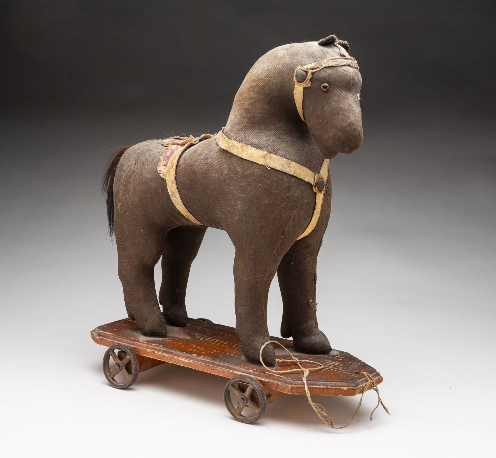 PENNSYLVANIA STRAW-STUFFED HORSE PULL TOY.: American, possibly Mennonite, Late 19th Century. Hand-stitched cloth horse with glass eyes, leather and faux-leather reins and saddle, and hair tail, on a shaped wooden base with iron wheels. 13"h. 5.