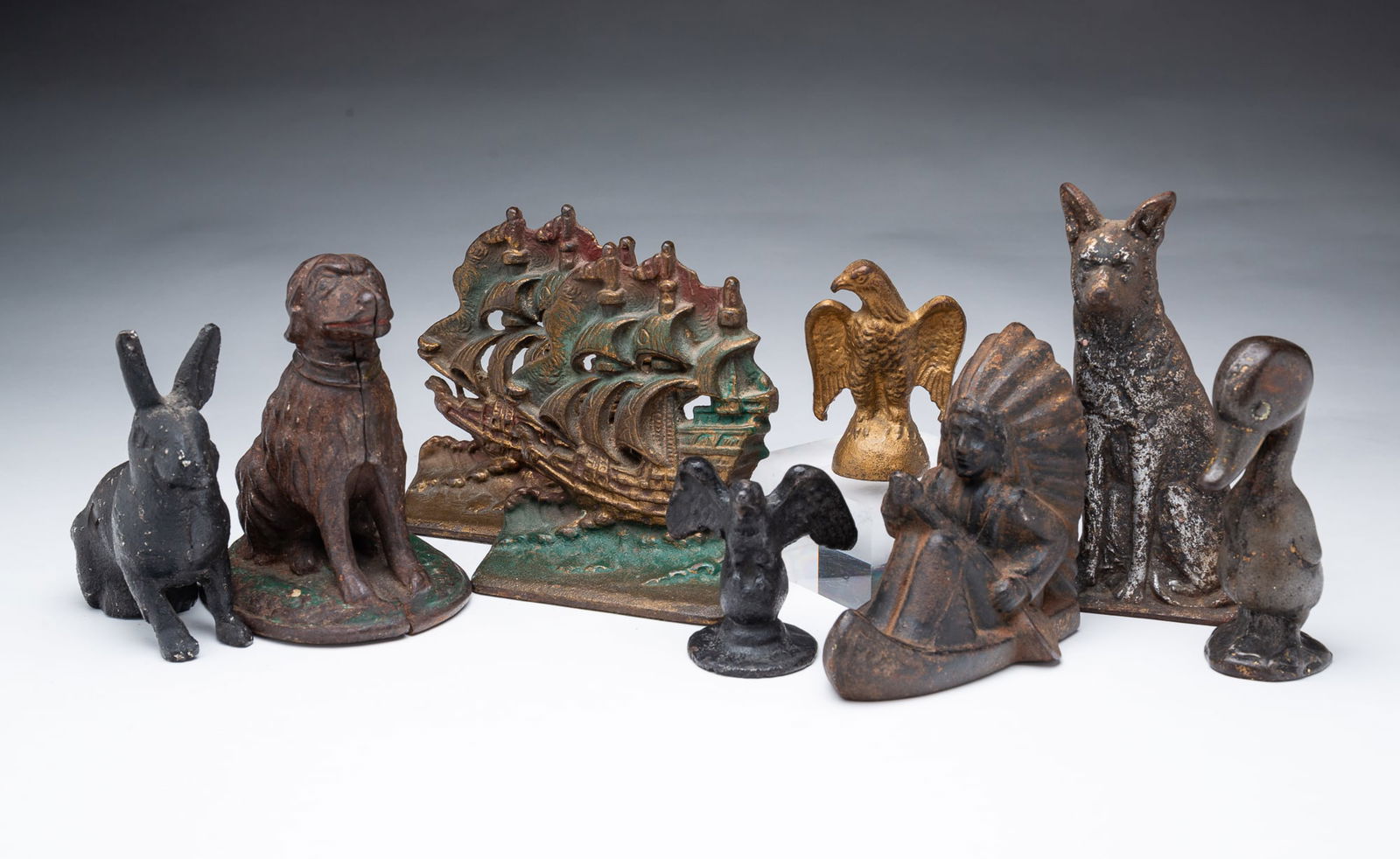 NINE AMERICAN CAST IRON FIGURES, INCL. GERMAN SHEPHERD DOORSTOP.: Ca. 1900. Comprising a bank of a Native American in a canoe, a pair of clipper ship bookends, a German Shepherd doorstop, a Labrador Retriever figure, a rabbit, two eagles, and a funny duck. Tallest,