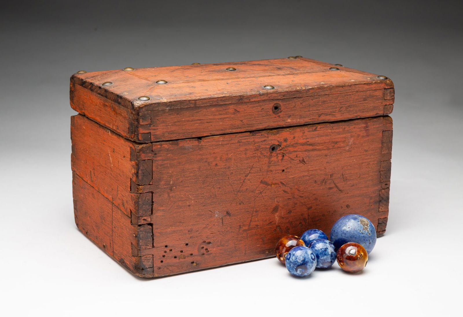 AMERICAN PINE BOX WITH HAND-ROLLED CLAY MARBLES.: Mid-19th Century. A pine box with dovetail construction, hinged lid decorated with brass tacks and in a red wash finish filled with hand rolled clay marbles in blue and brown. 5.75"h. 9.5"d. 6"d.Prove