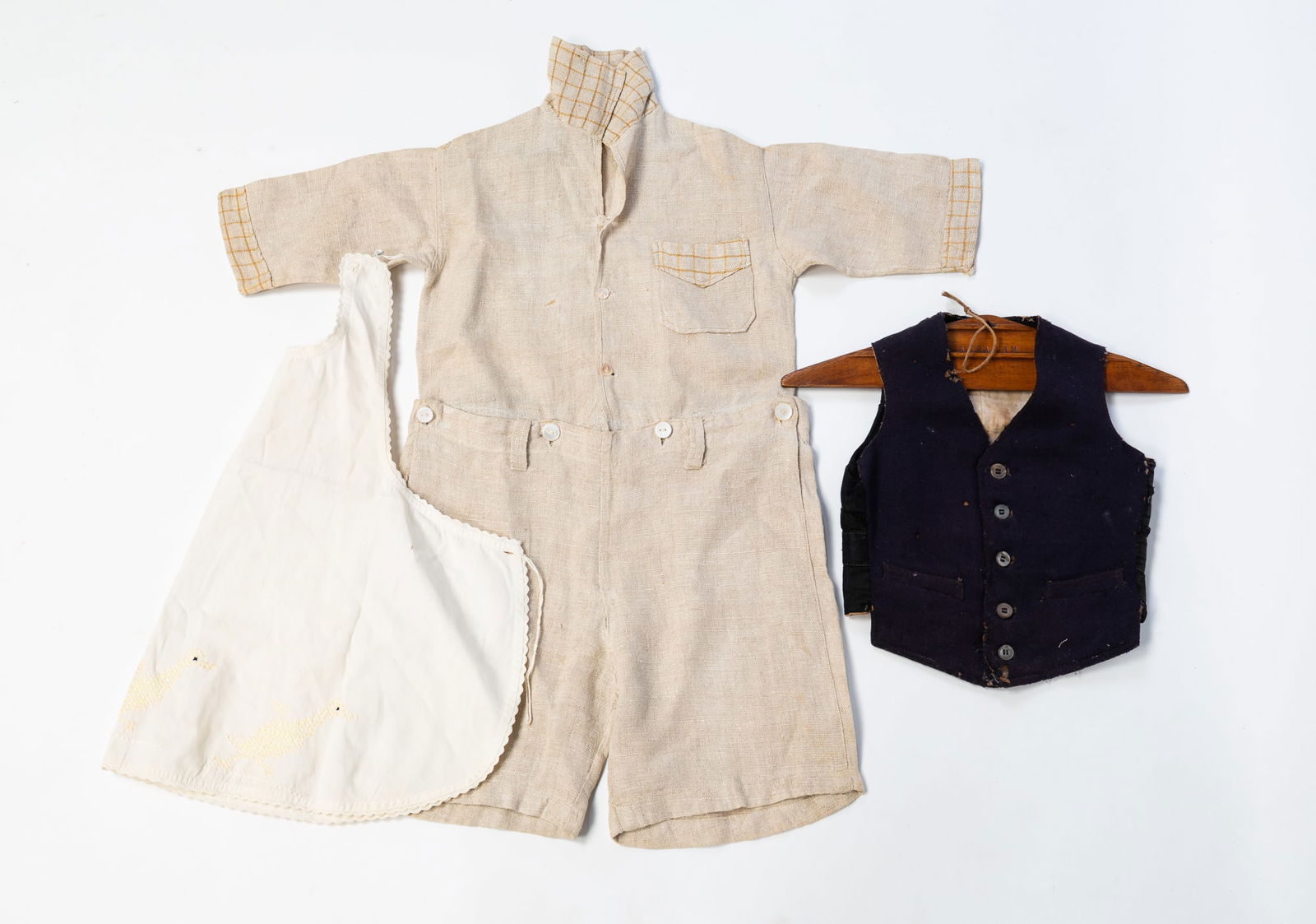 THREE ARTICLES OF CHILDREN'S CLOTHING.: American, Early-mid 20th Century. Machine-sewn. Homespun linen romper with collar, pocket, and buttons, blue wool and cotton vest with a wooden hanger, cotton bib apron cross-stitched with ducks. In v